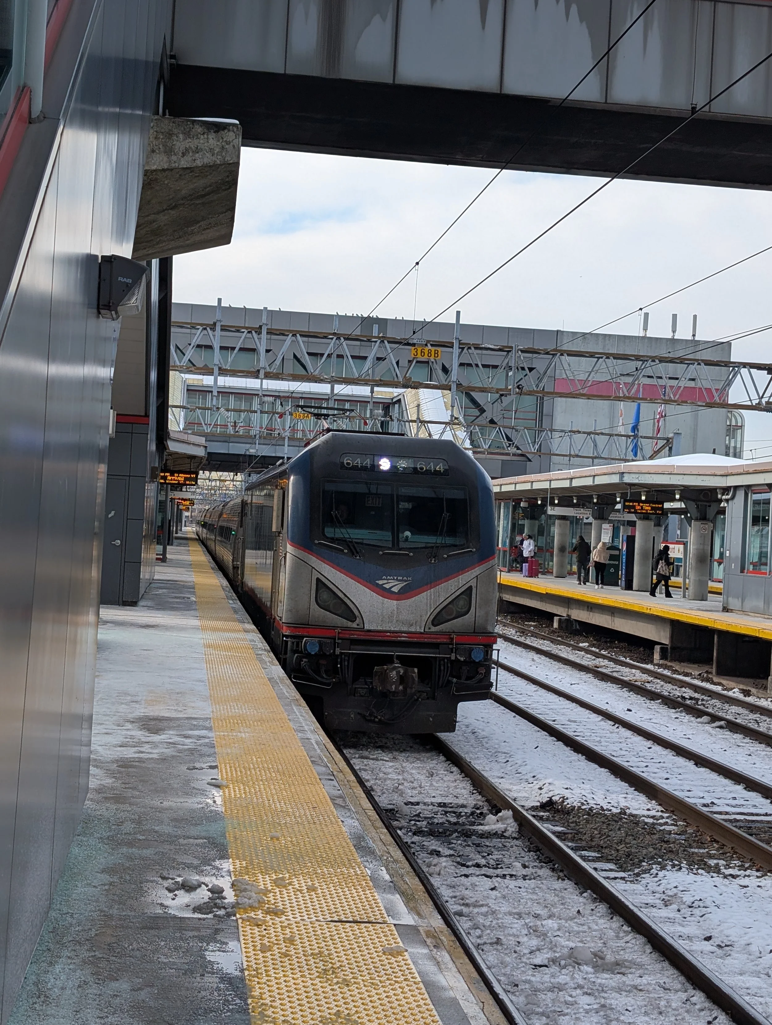 A black and gray Amtrak train at a modern outdoor train station with snow on the platform and tracks, and a few passengers waiting.