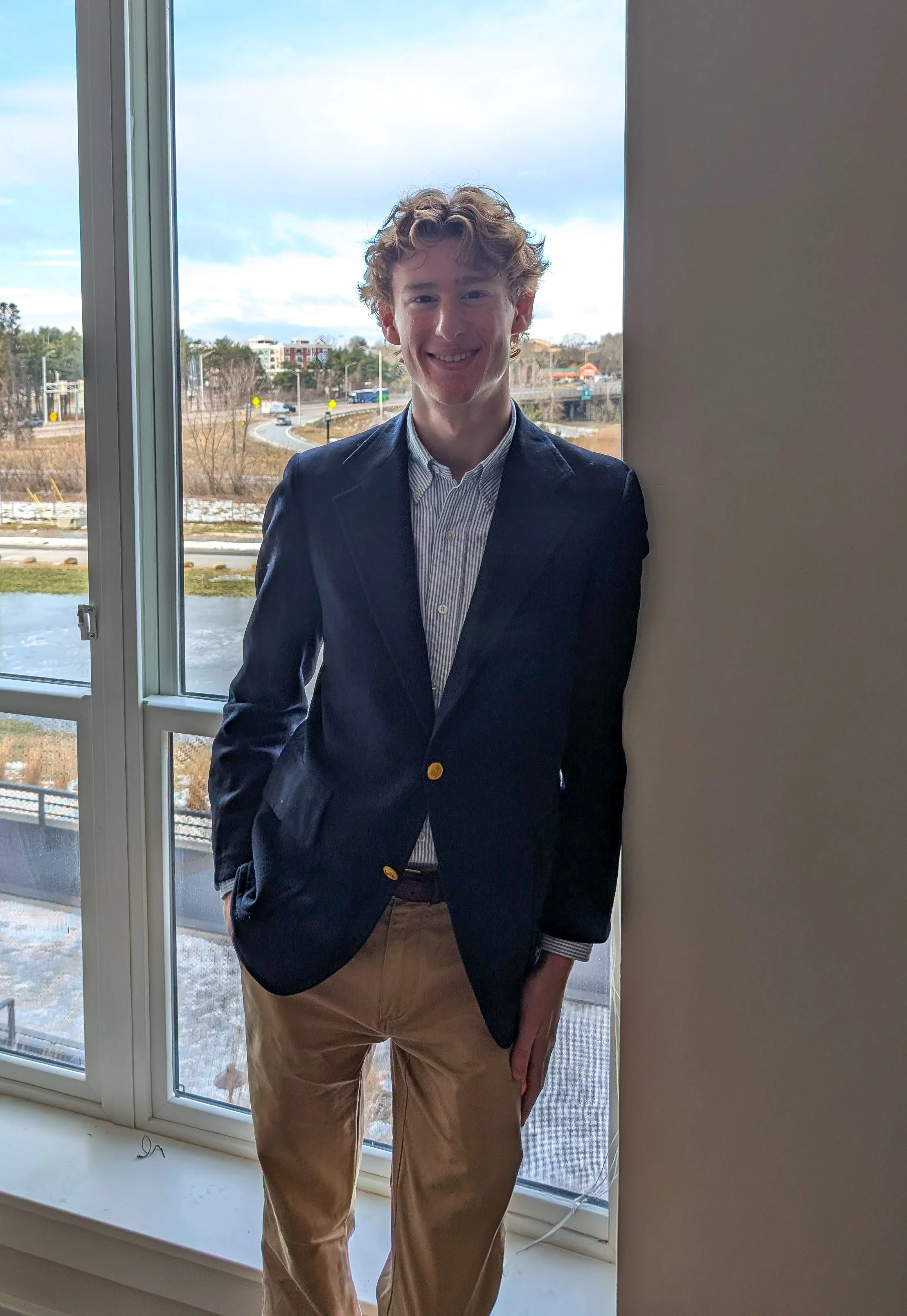 A young man with curly red hair wearing a navy blazer, striped shirt, and khaki pants standing next to a window with a view of a winter landscape and a street, smiling at the camera.