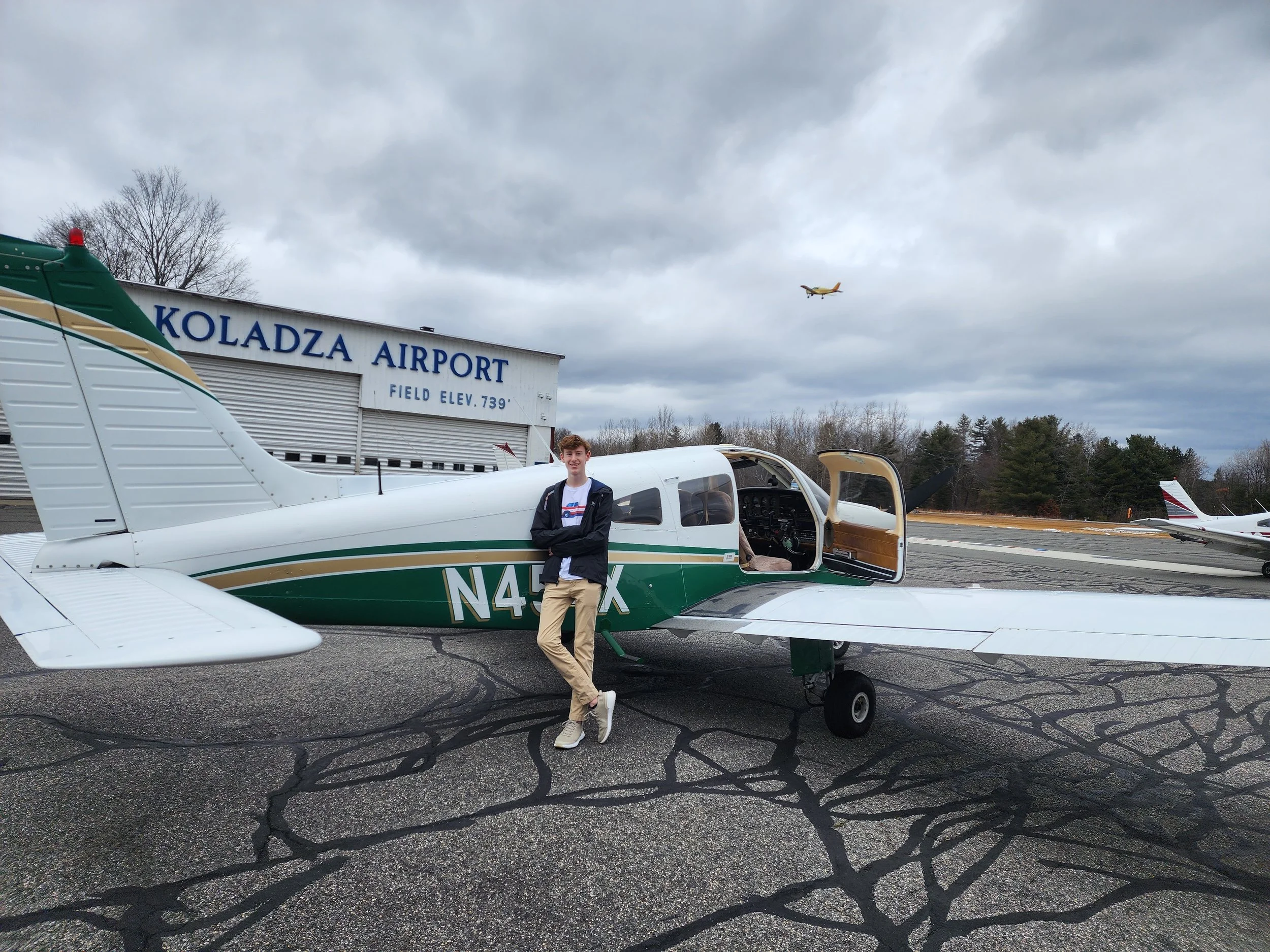 A young man in beige pants and a dark jacket standing next to a small green and white airplane parked outside Koladza Airport. The airport sign is visible in the background, along with a few other small planes and a cloudy sky with an airplane flying overhead.