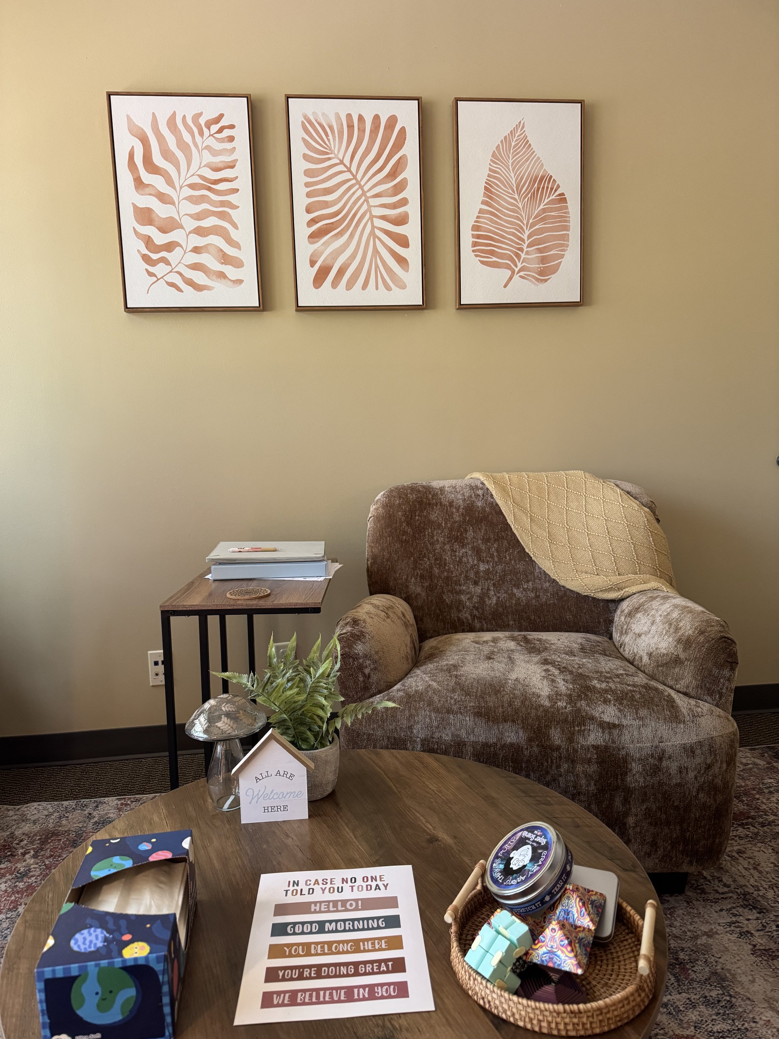 A cozy living room corner with a brown armchair, a wooden side table, and a round wooden table. The wall has three framed leaf art prints. The round table has a vase with green plants, a sign saying "All are Welcome Here," and a colorful card box. The main table also has a sheet with positive messages and some game pieces.
