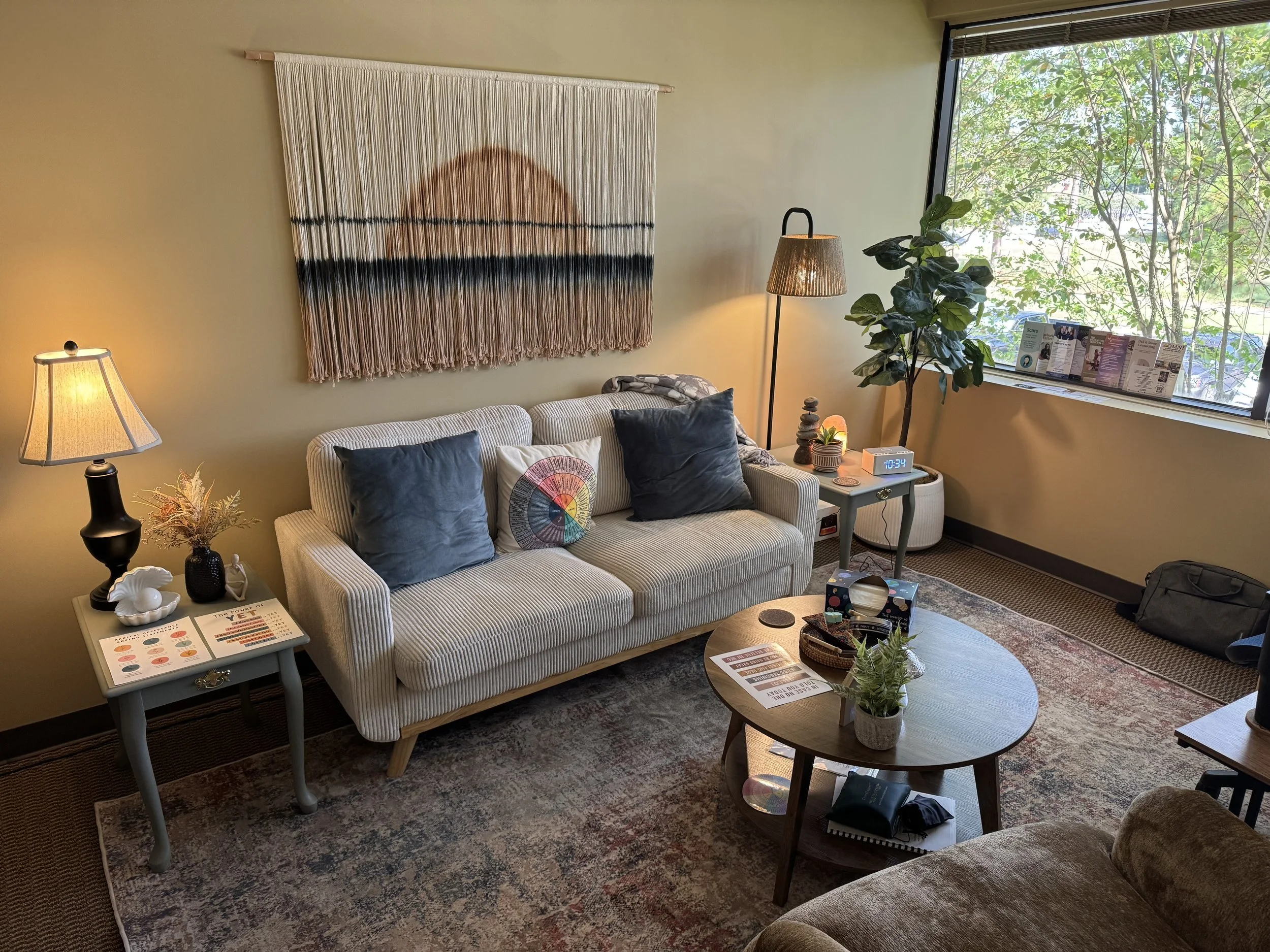 Cozy living room with beige walls, a white striped sofa with cushions, a side table with a lamp, a floor lamp, a large window with greenery outside, a potted plant near the window, and a coffee table with plants and papers.