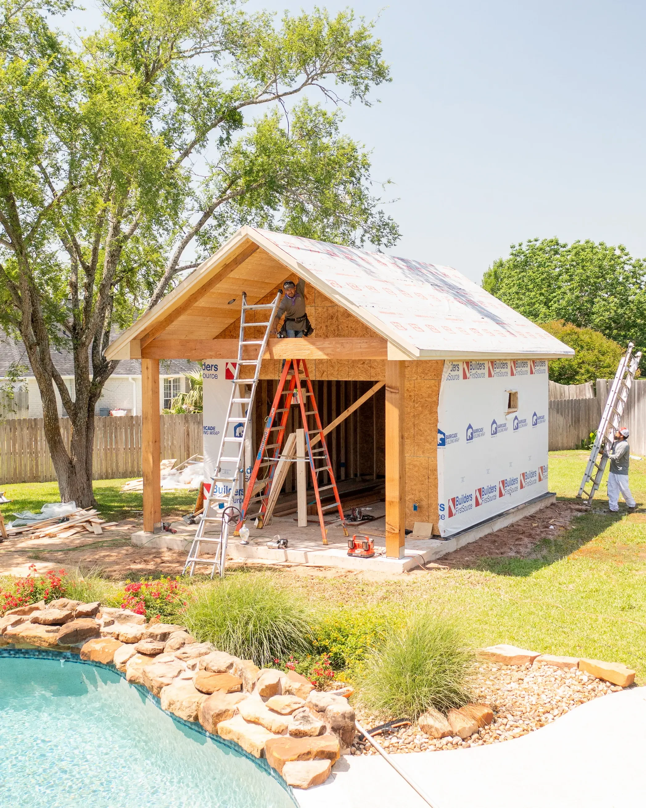 A construction worker on a ladder working on a house frame with a roof, surrounded by trees, a fenced backyard, and a swimming pool in the foreground.