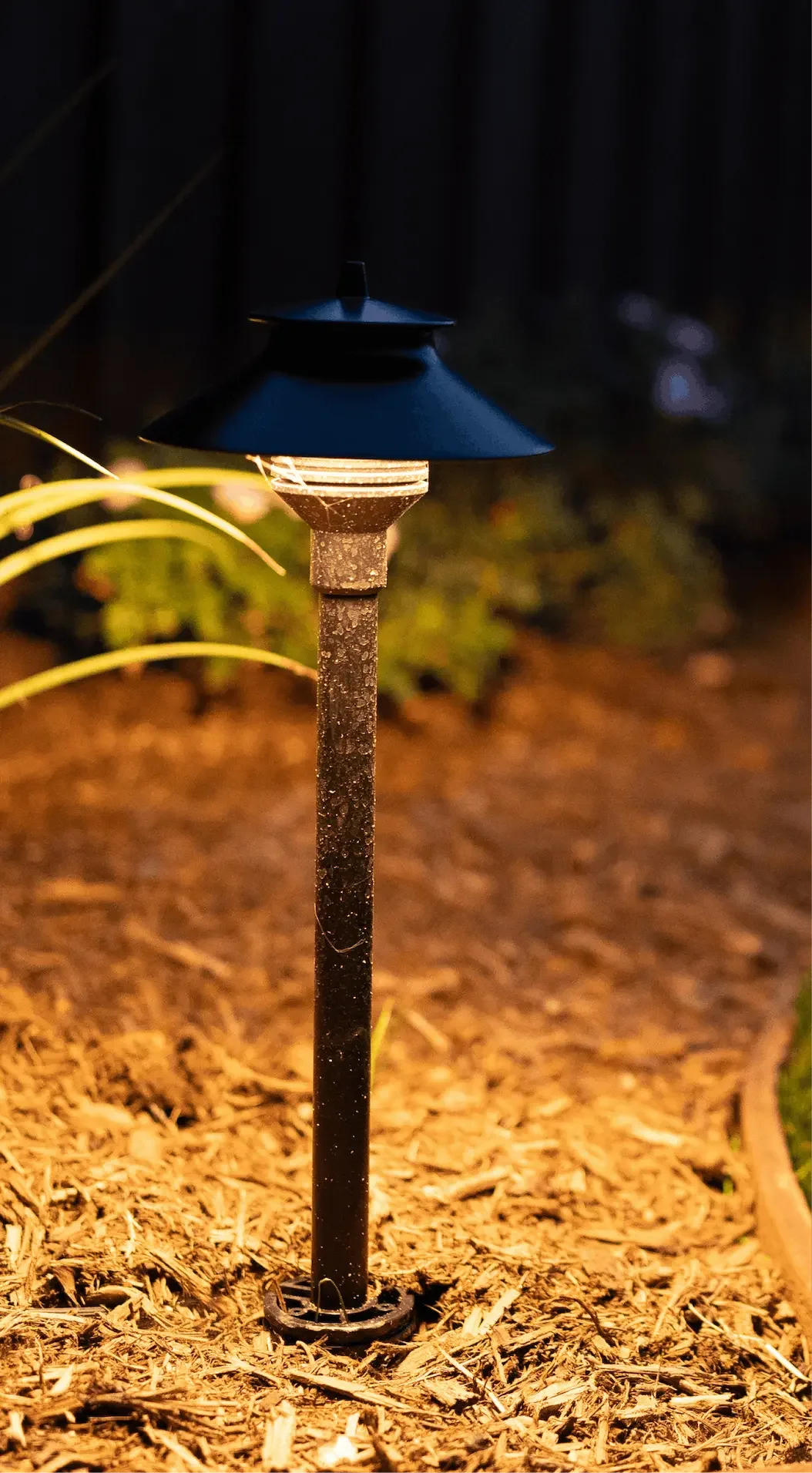 A black outdoor solar garden light with a decorative top, installed in soil surrounded by mulch, emitting a warm glow at night.
