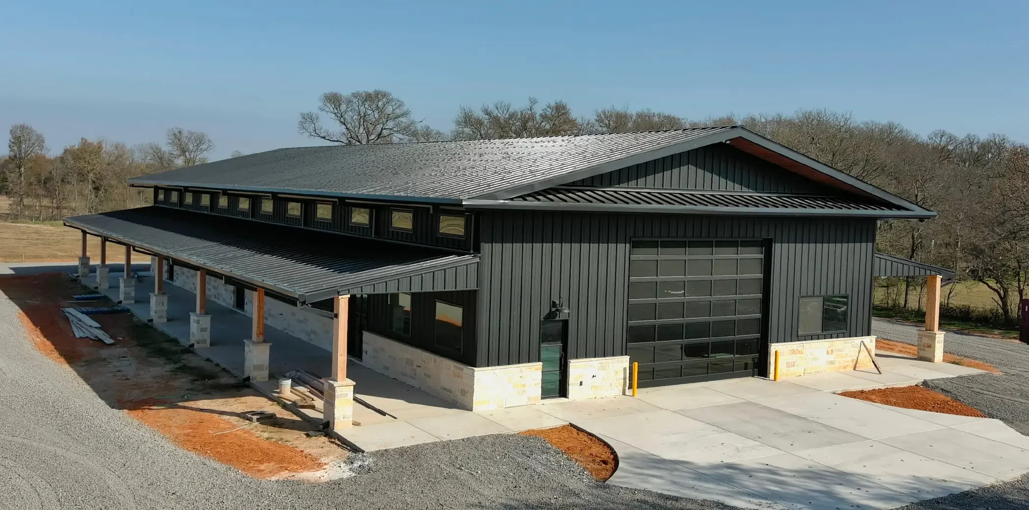 Newly constructed building with black metal siding, large glass garage door, and a covered porch area, set in a suburban area with trees in the background.