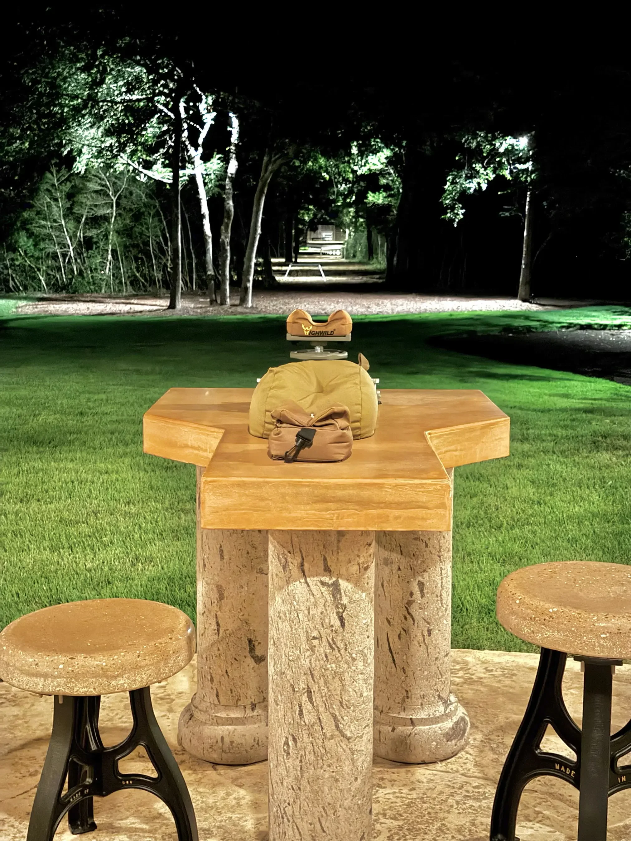 Indoor scene with a marble table, three round seats, and a tall structure in the middle, set against a backdrop of a dark wooded area with trees and uneven ground.