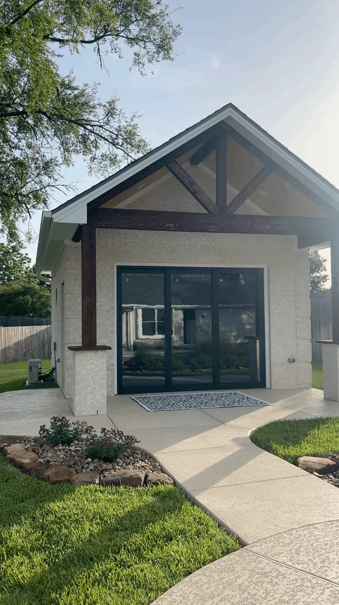 The image shows the back of a house with a small covered patio, black sliding glass doors, a patterned outdoor rug, and well-maintained yard with green grass, edging stones, and a concrete walkway.