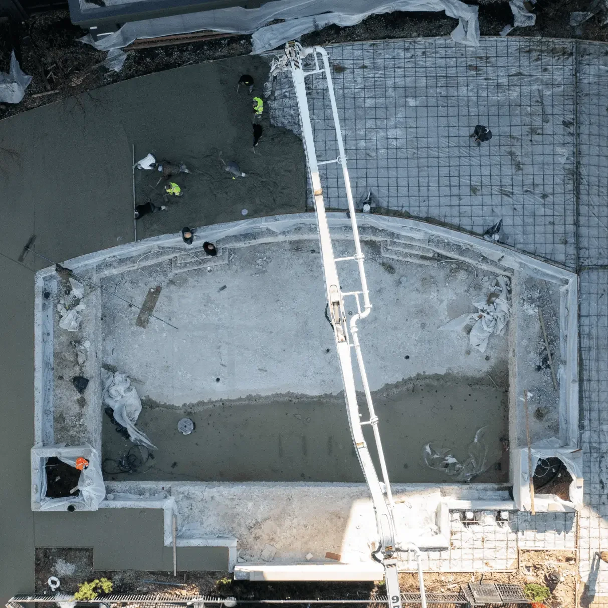 Aerial view of a construction site with workers pouring concrete into a foundation or pool, with gravel and rebar grids on the ground surrounding the area.