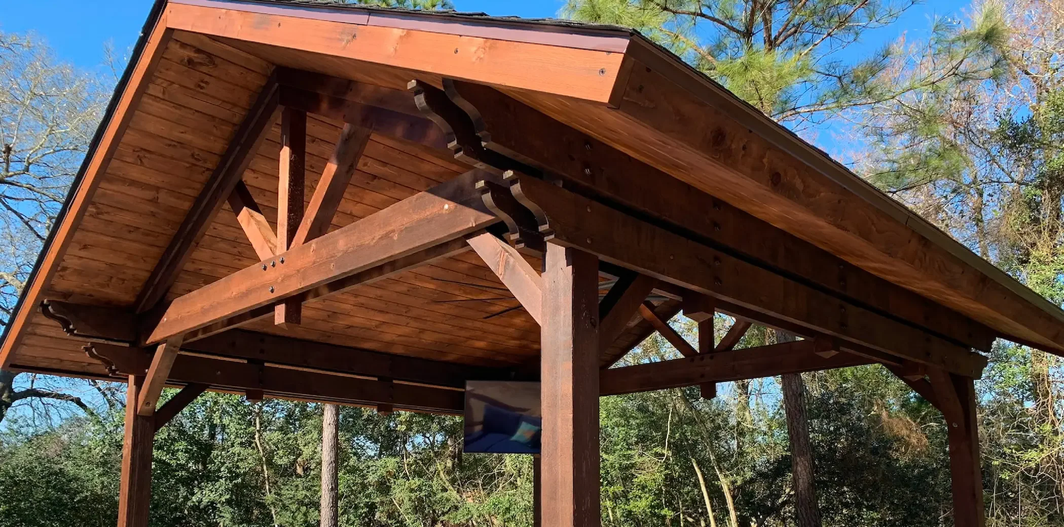 View of a wooden outdoor pavilion or gazebo with a sloped roof, supported by wooden beams, in a wooded area with trees and clear blue sky in the background.