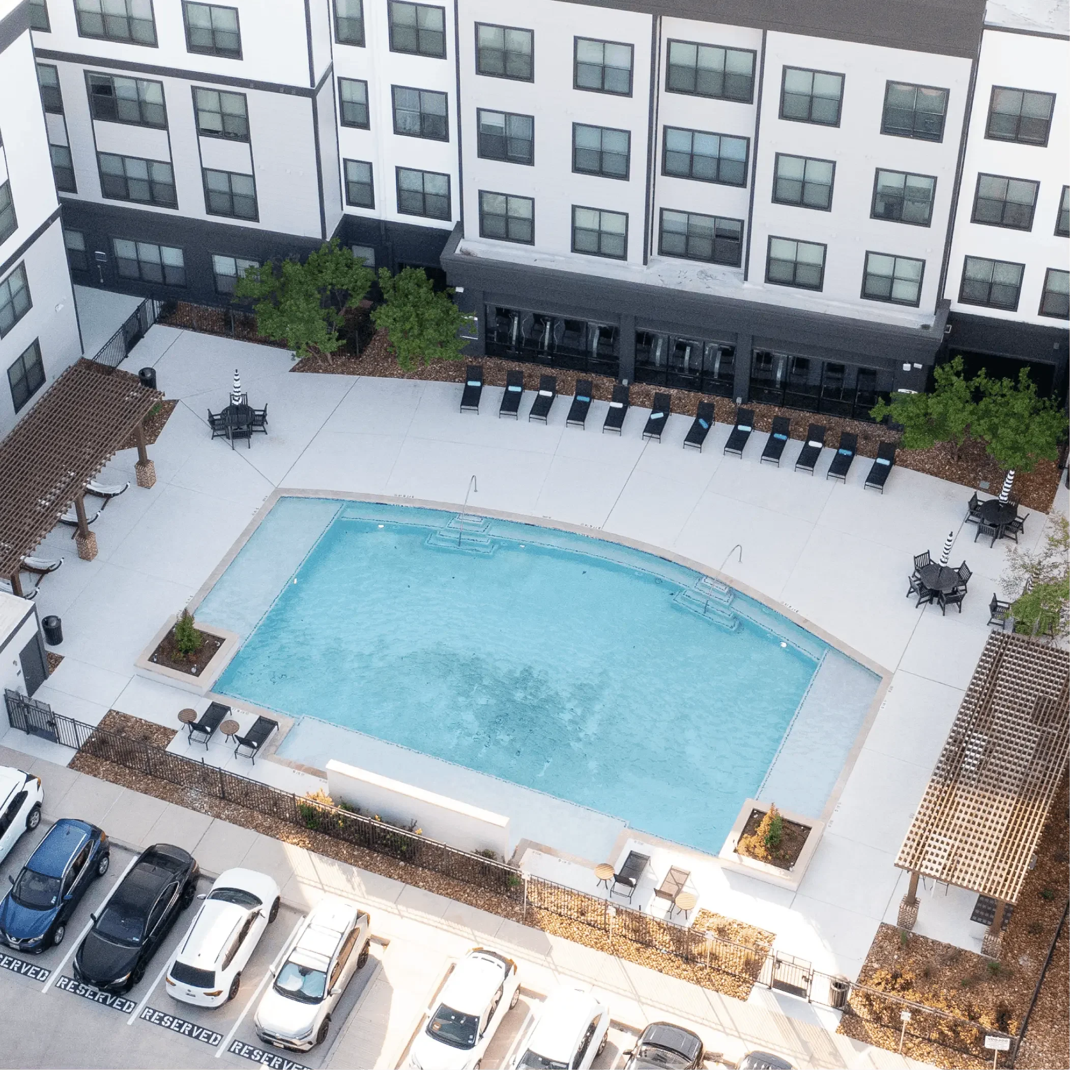 A view of a multi-story apartment building with a parking lot in front. The parking lot has cars parked in designated spaces labeled 'RESERVED'. The building has multiple windows and a small outdoor area with chairs and tables, a tree, and a swimming pool.