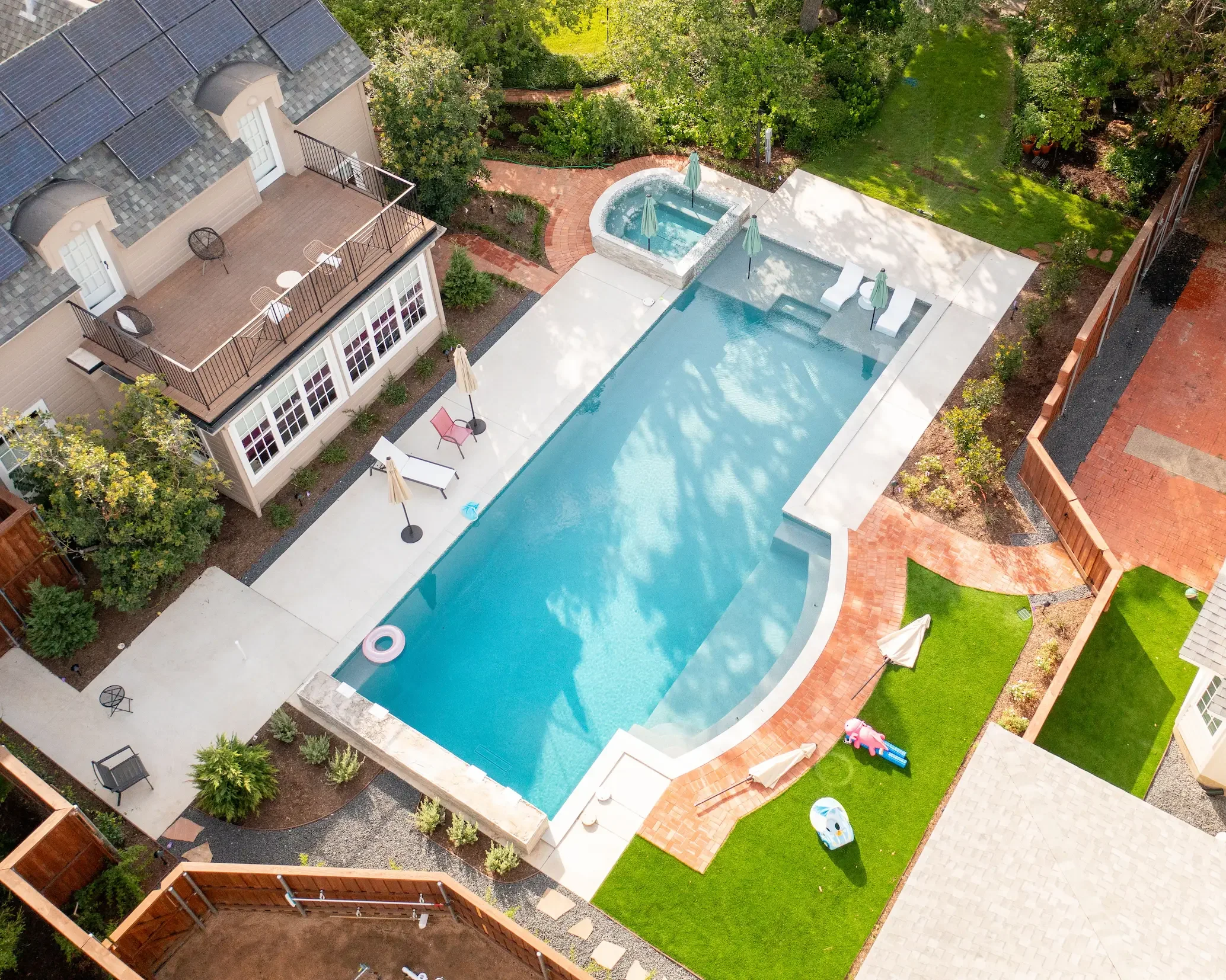 An aerial view of a backyard with a swimming pool, hot tub, outdoor furniture, a deck, a grassy lawn, trees, and a brick pathway in a suburban neighborhood.