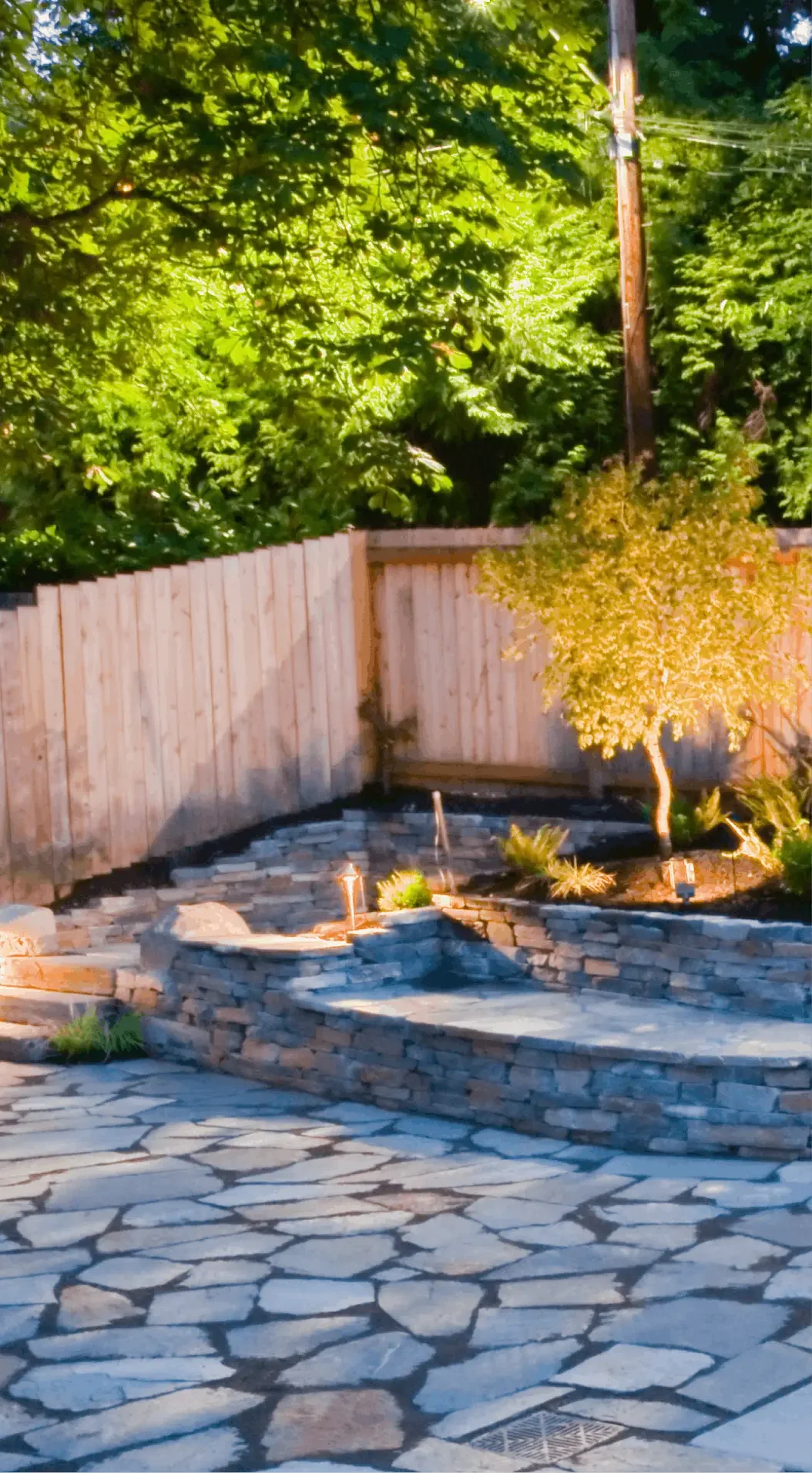 A backyard with a stone patio, a small water fountain, a wooden fence, and green trees, captured during early evening with sunlight.