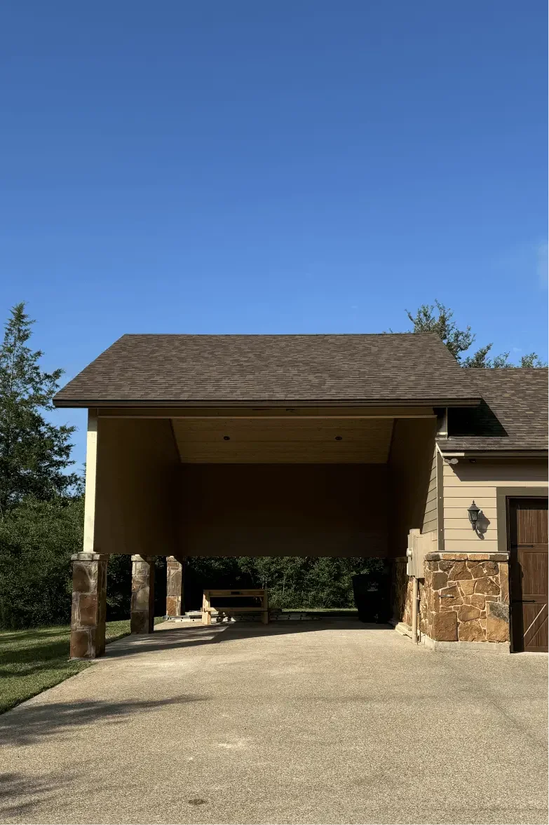 A house with a carport supported by stone and wood columns, with a gravel driveway and a wooden bench underneath, against a background of trees and a blue sky.