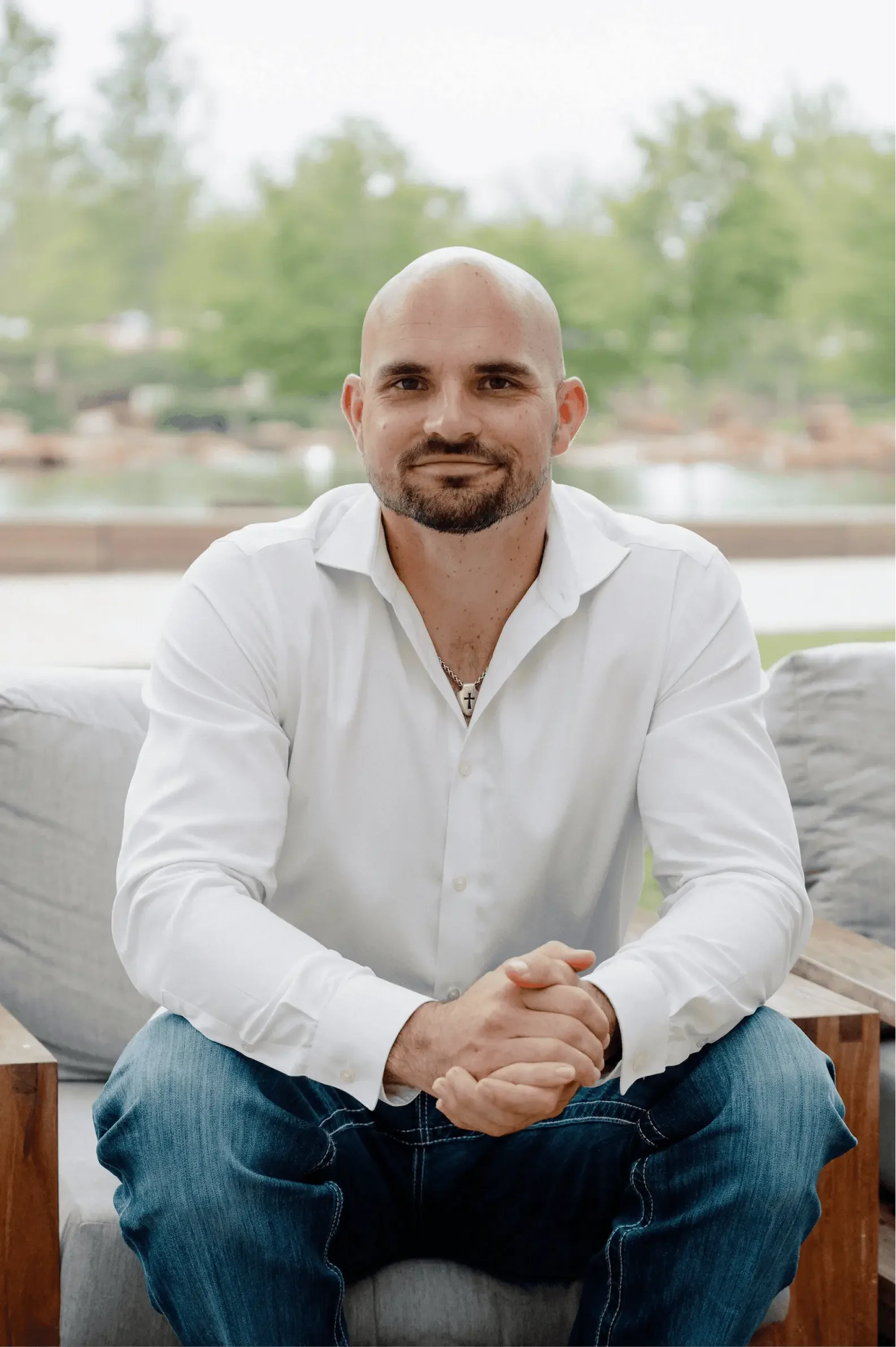 A man with a bald head, beard, and mustache, wearing a white button-up shirt and blue jeans, sitting on a sofa with his hands clasped, smiling slightly, in front of a window with a blurry green outdoor background.