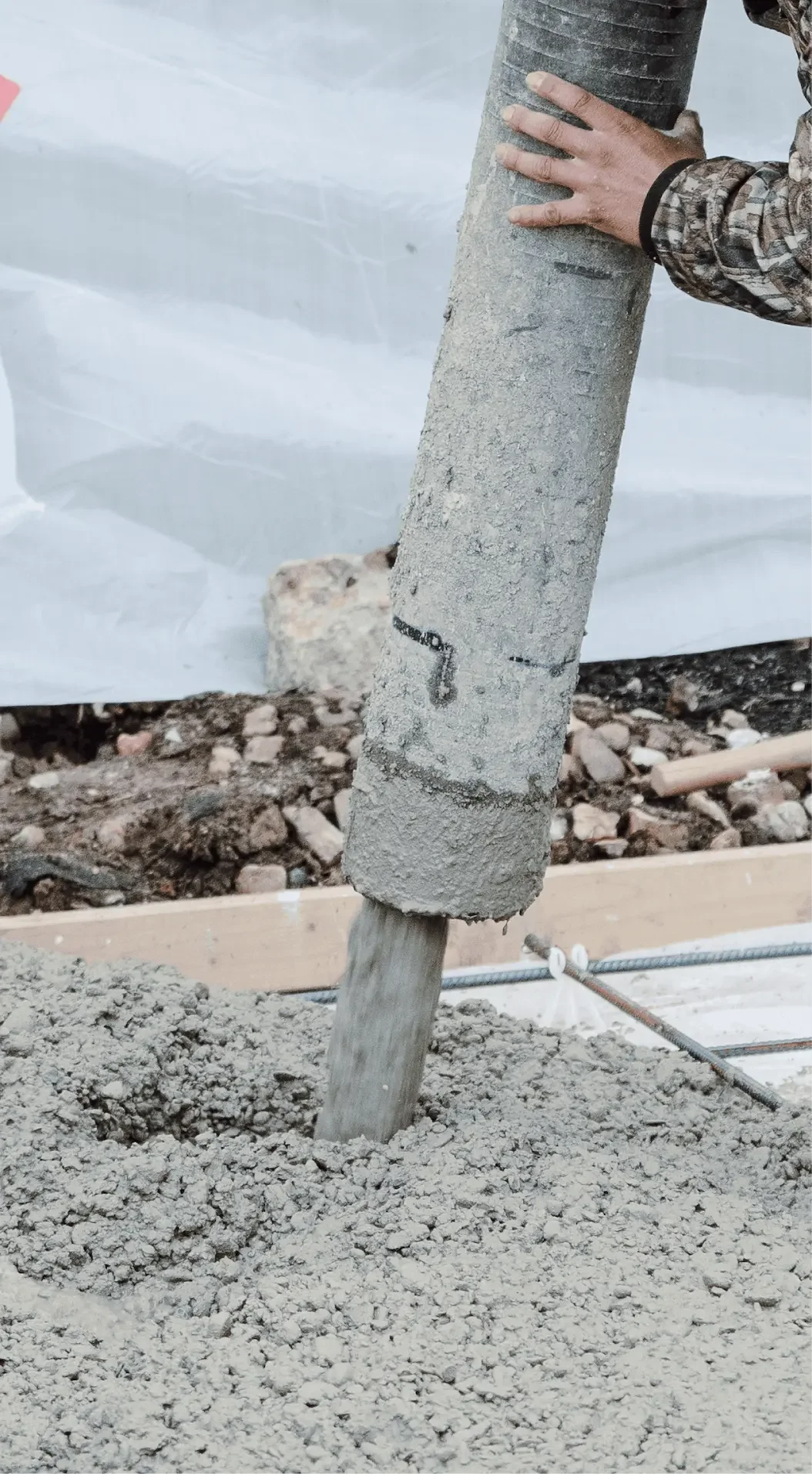 A worker installs a concrete pole into the ground at a construction site. The pole is partially embedded in the soil, with one hand guiding it into place.