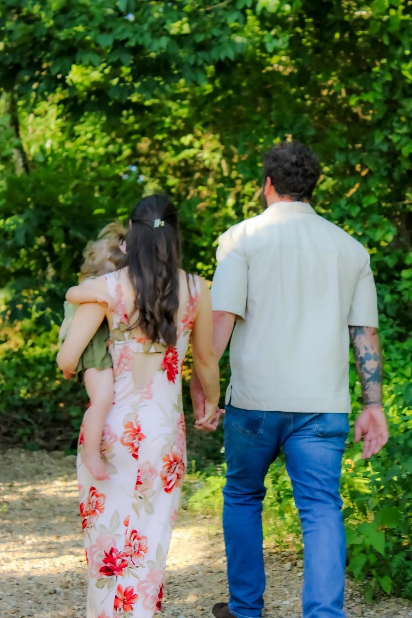 A couple holding hands and walking with a young girl in a floral dress, outdoors with green foliage in the background.