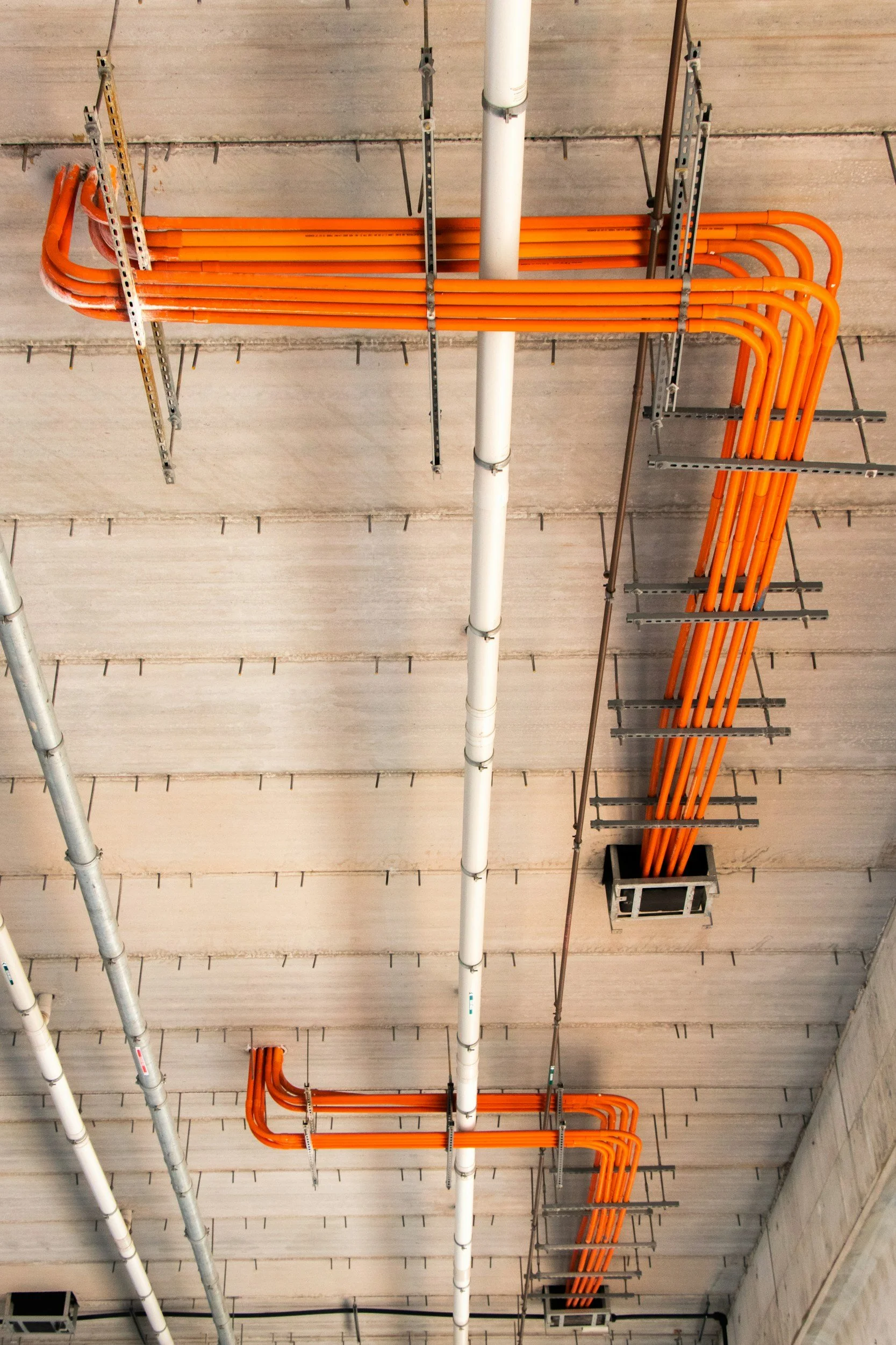 Ceiling with white, gray, and orange pipes and ductwork, supported by metal brackets, against a beige wall.
