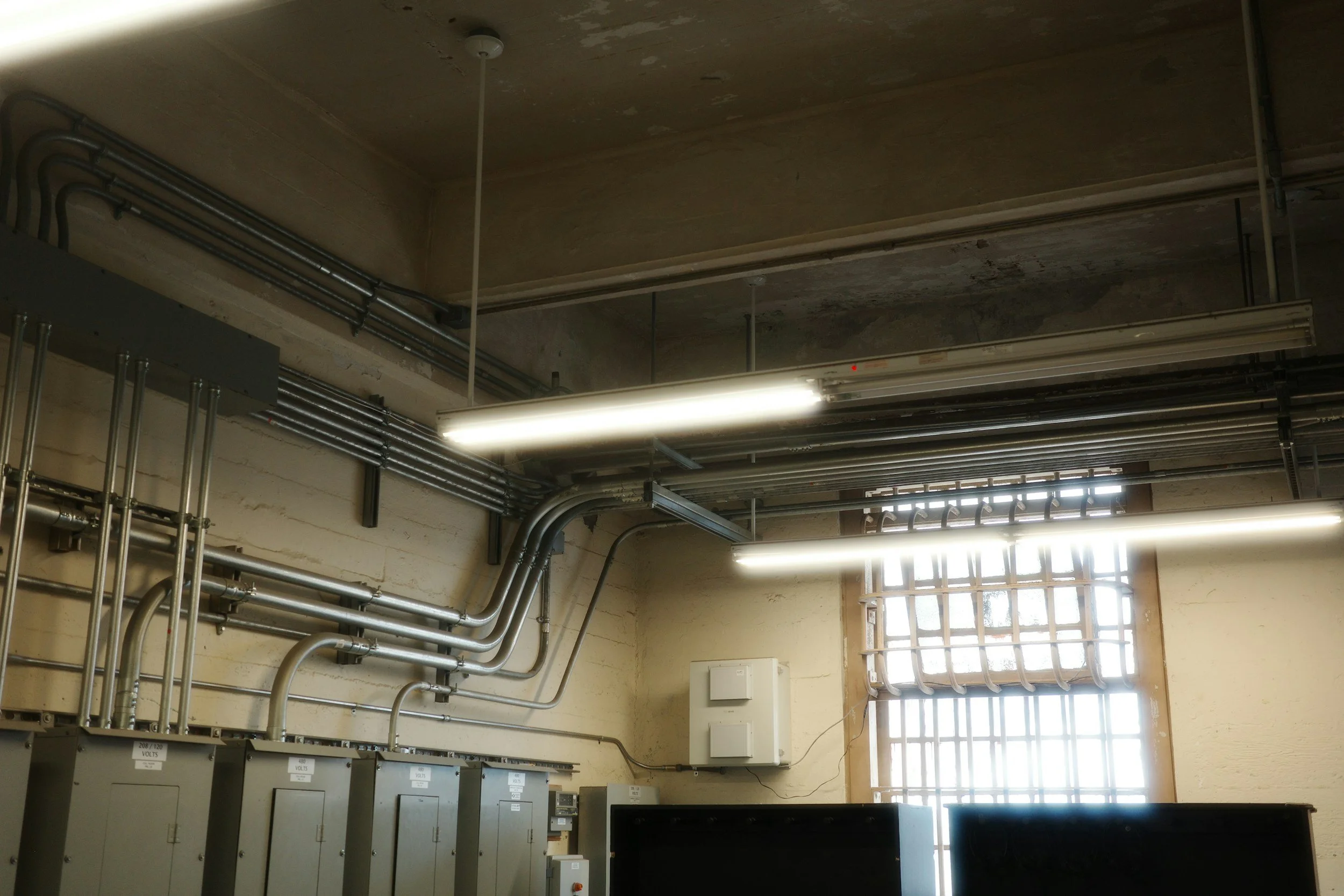 Electrical wiring, pipes, and electrical panels in an industrial or utility room with a window allowing natural light.