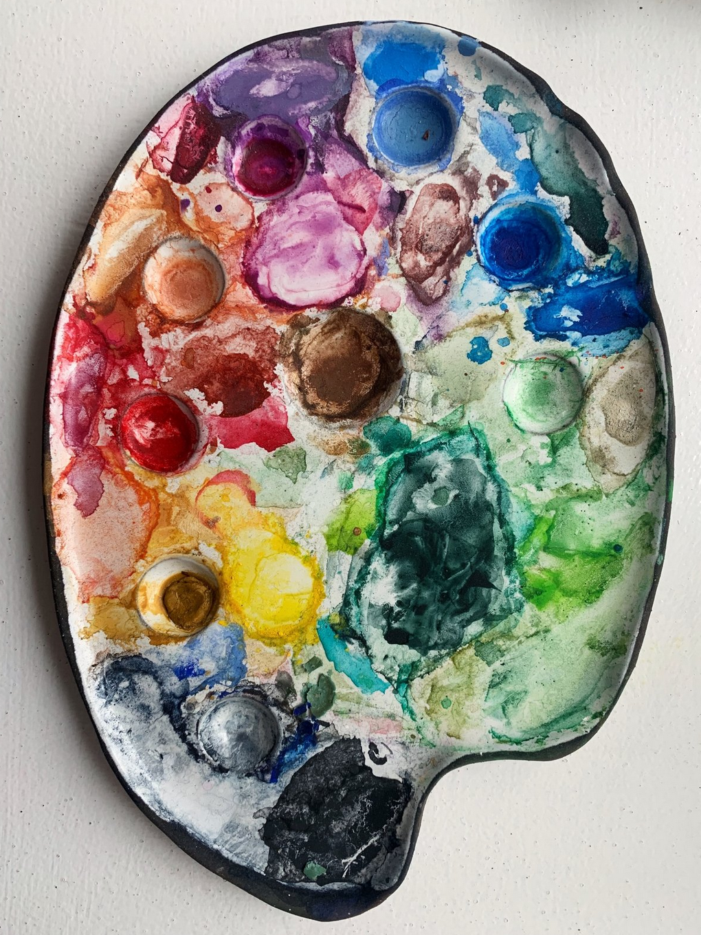A watercolor paint palette with multiple wells filled with different colors, including purple, blue, green, yellow, red, and brown, used for painting.