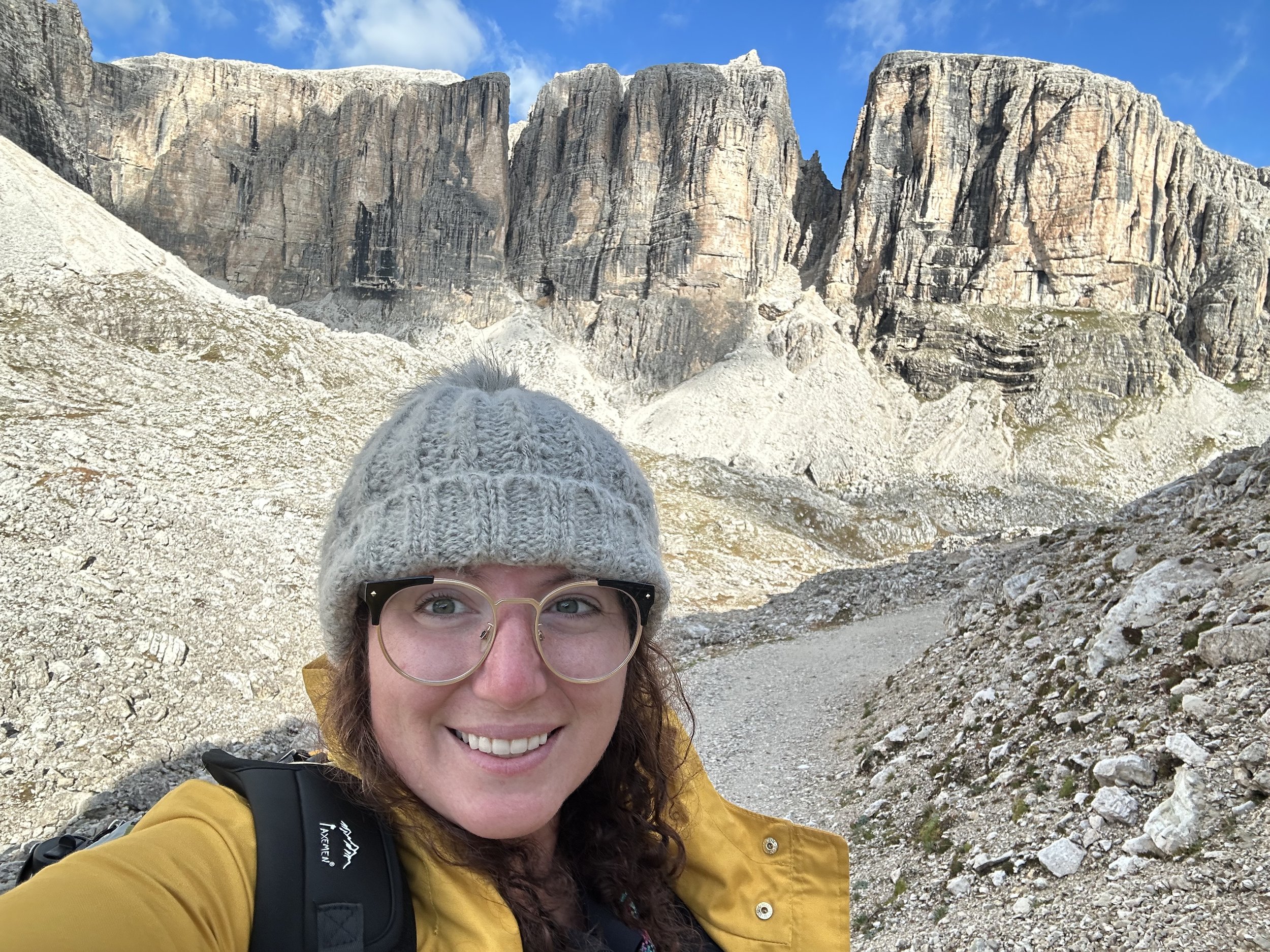 A woman wearing glasses, a gray knit beanie, and a yellow jacket taking a selfie in a rocky mountain landscape with towering cliffs and a blue sky in the background.