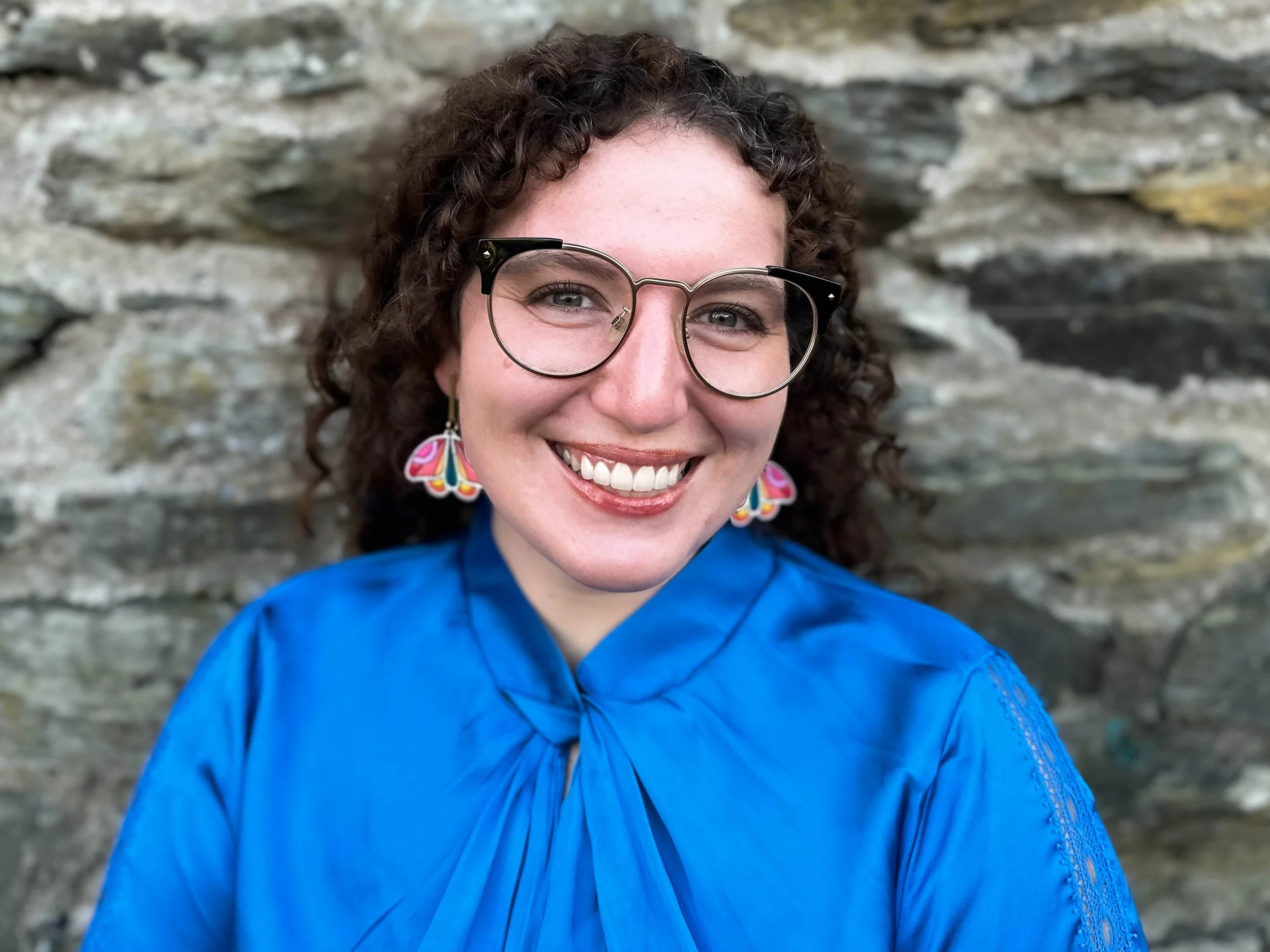 A woman with curly brown hair and glasses smiling, wearing a bright blue top and colorful butterfly earrings, standing in front of a rocky background.