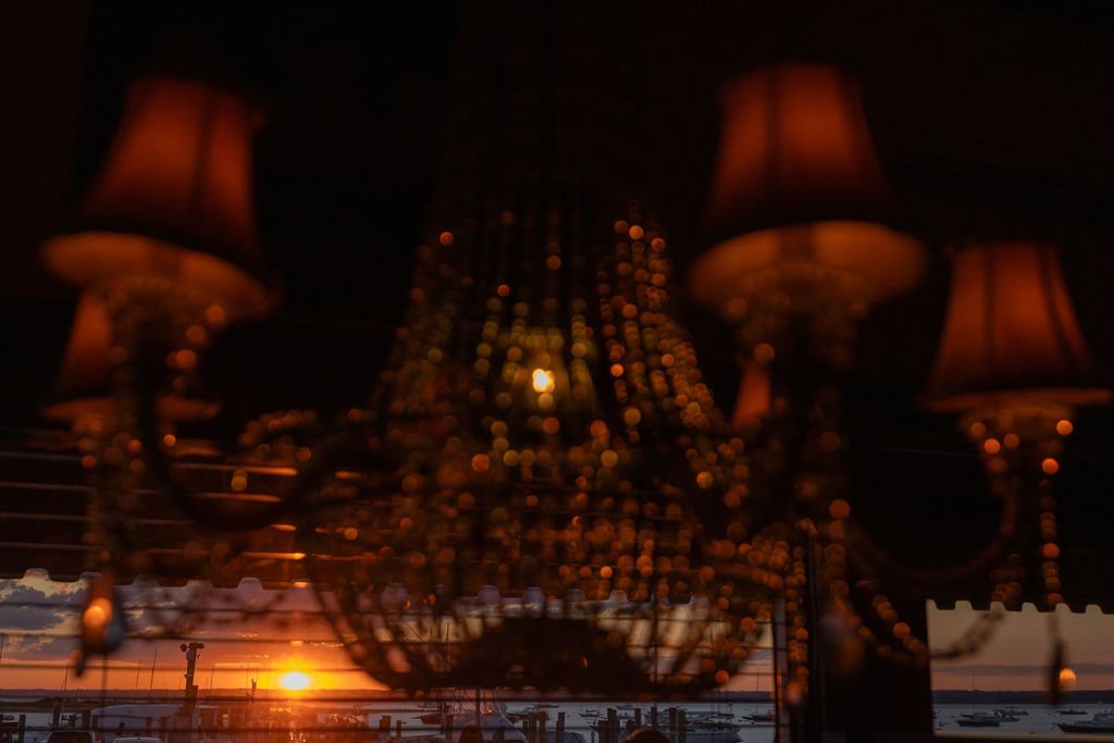 A chandelier with multiple lampshades and hanging beads, reflecting a sunset view with boats at the water's edge.