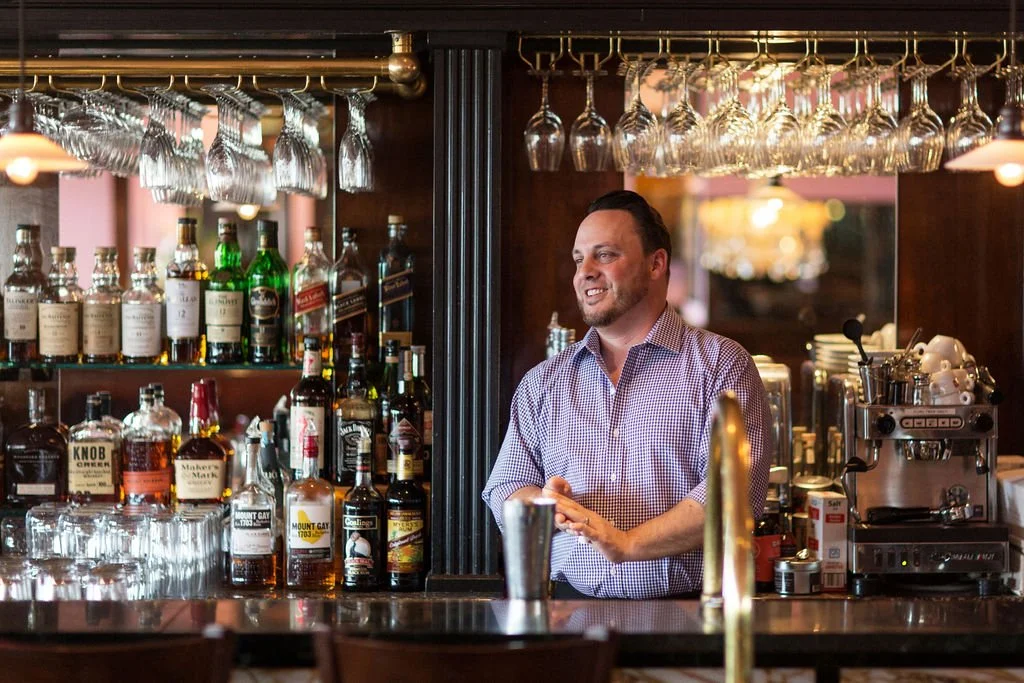 A bartender standing behind a bar counter, smiling, with various liquor bottles and glassware hanging above him in a bar or restaurant setting.