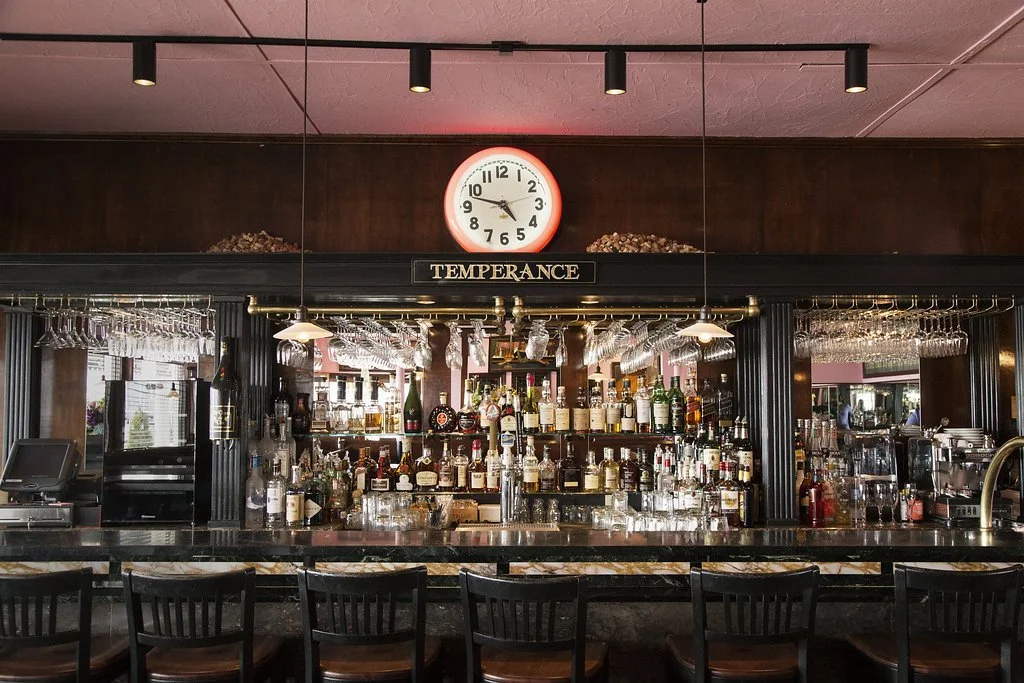 Bar with shelves of alcohol bottles, hanging glasses, and a clock showing 10:10, with a sign labeled 'Temperance' above the bar.