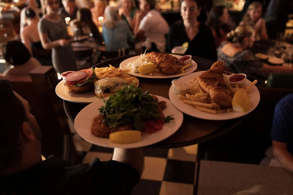 A waiter serving plates of food, including sandwiches, fried chicken with fries, and a salad, in a dimly lit restaurant with customers dining in the background.