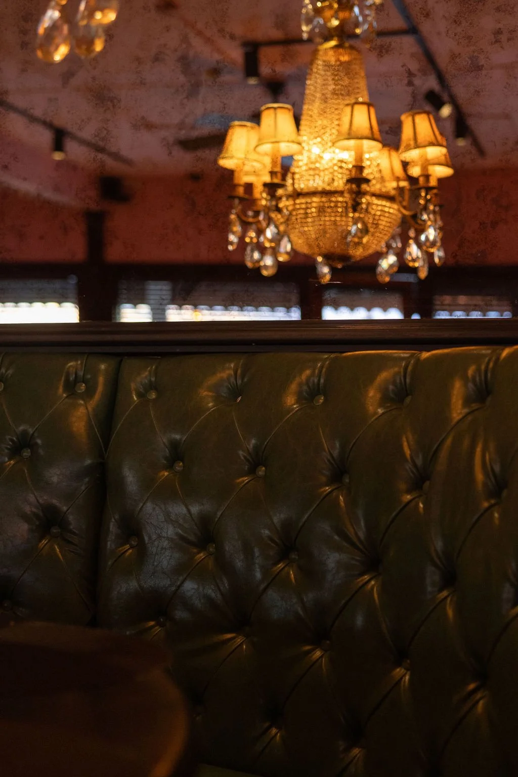 Chandelier hanging from a ceiling above a tufted leather booth in a dimly lit restaurant or bar.