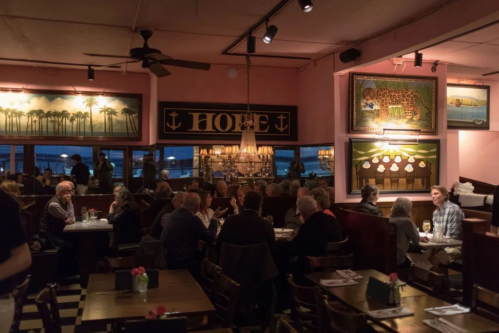 A lively restaurant with people dining, colorful artwork on the walls, and a chandelier hanging from the ceiling.