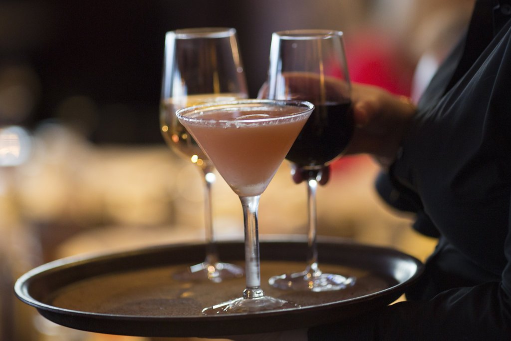 A server holding a tray with three glasses of wine and a cocktail in a dimly lit bar or restaurant.