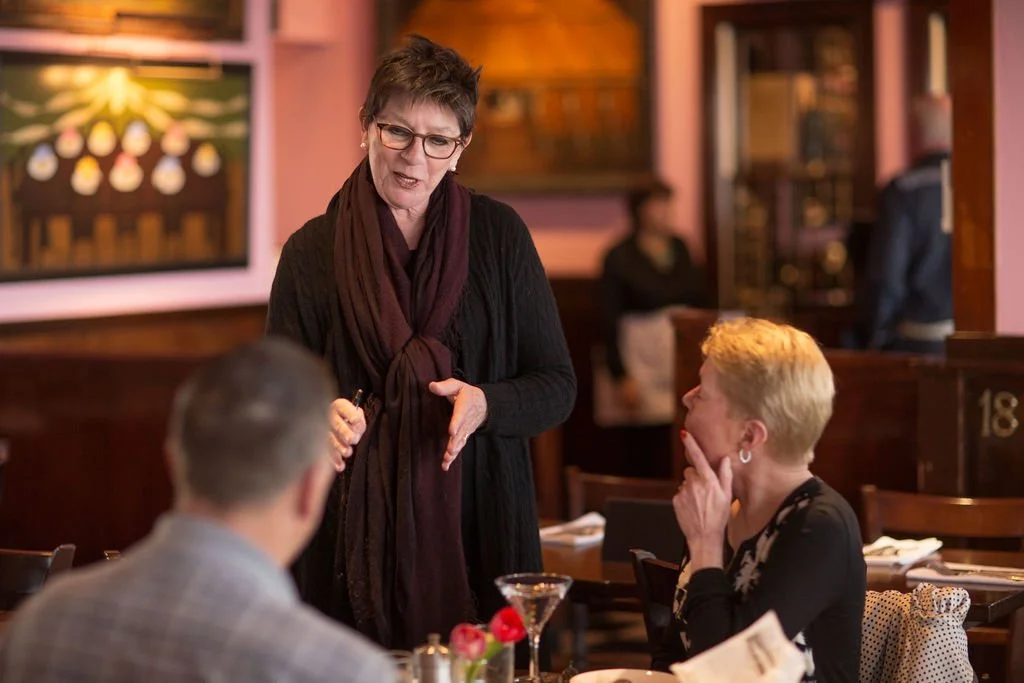 A woman with short dark hair, glasses, and a dark scarf is speaking to a man and a woman at a restaurant table. The woman has short blonde hair, and the man has short brown hair. The table has a martini glass, a small flower arrangement, and menus. T