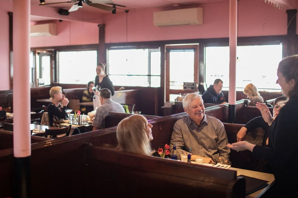 People dining at a restaurant, smiling and engaging in conversation, with a waitress serving a table, and large windows letting in natural light.
