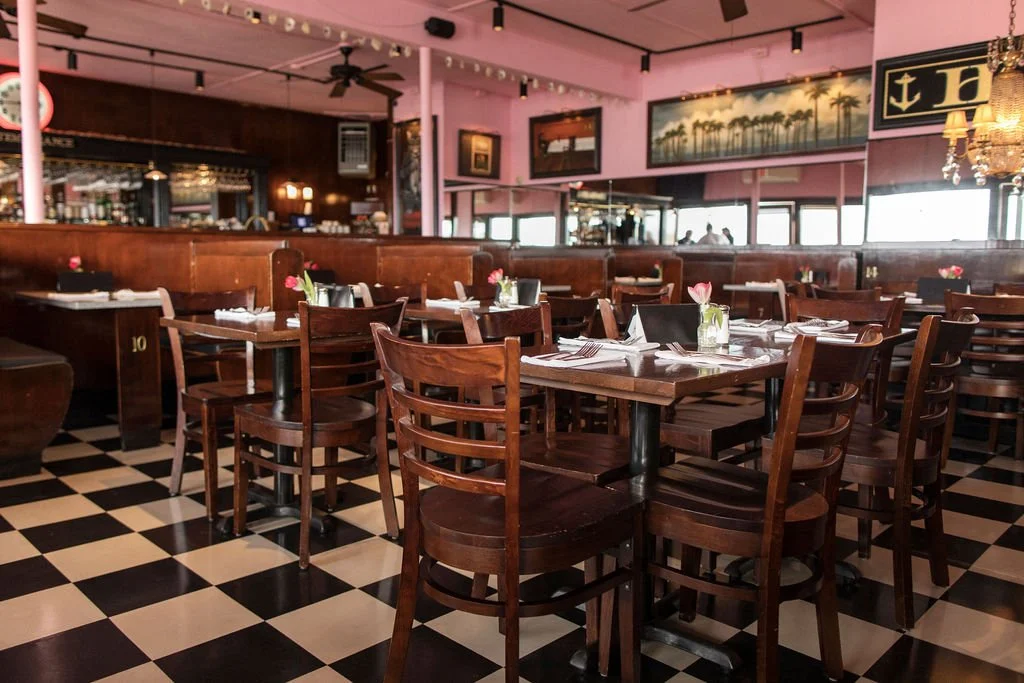 Interior of a restaurant with wooden tables and chairs, pink walls decorated with paintings, and a black and white checkered floor.