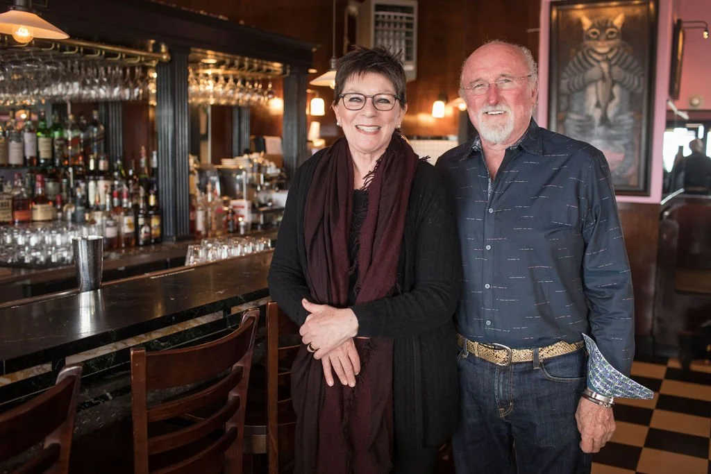 A smiling woman with short dark hair and glasses, wearing a black outfit with a dark red scarf, standing next to a smiling man with glasses, a white beard, wearing a dark blue shirt, in a dimly lit bar or restaurant with a bar counter, bottles, glass