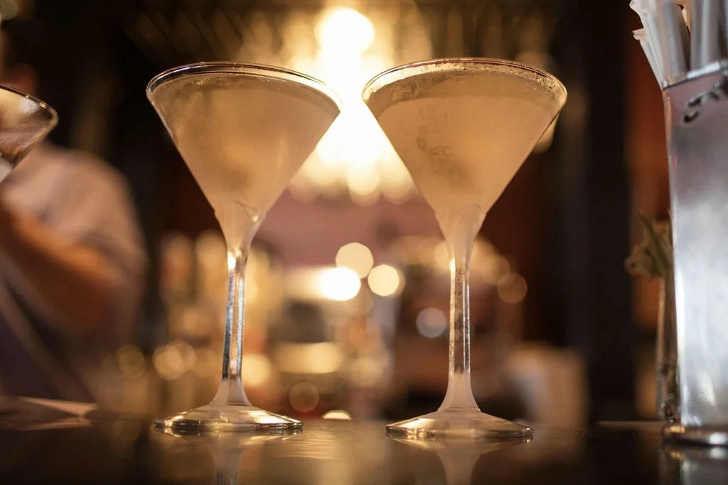 Two cocktails in martini glasses with a creamy pale color on a bar counter, with warm lighting in the background