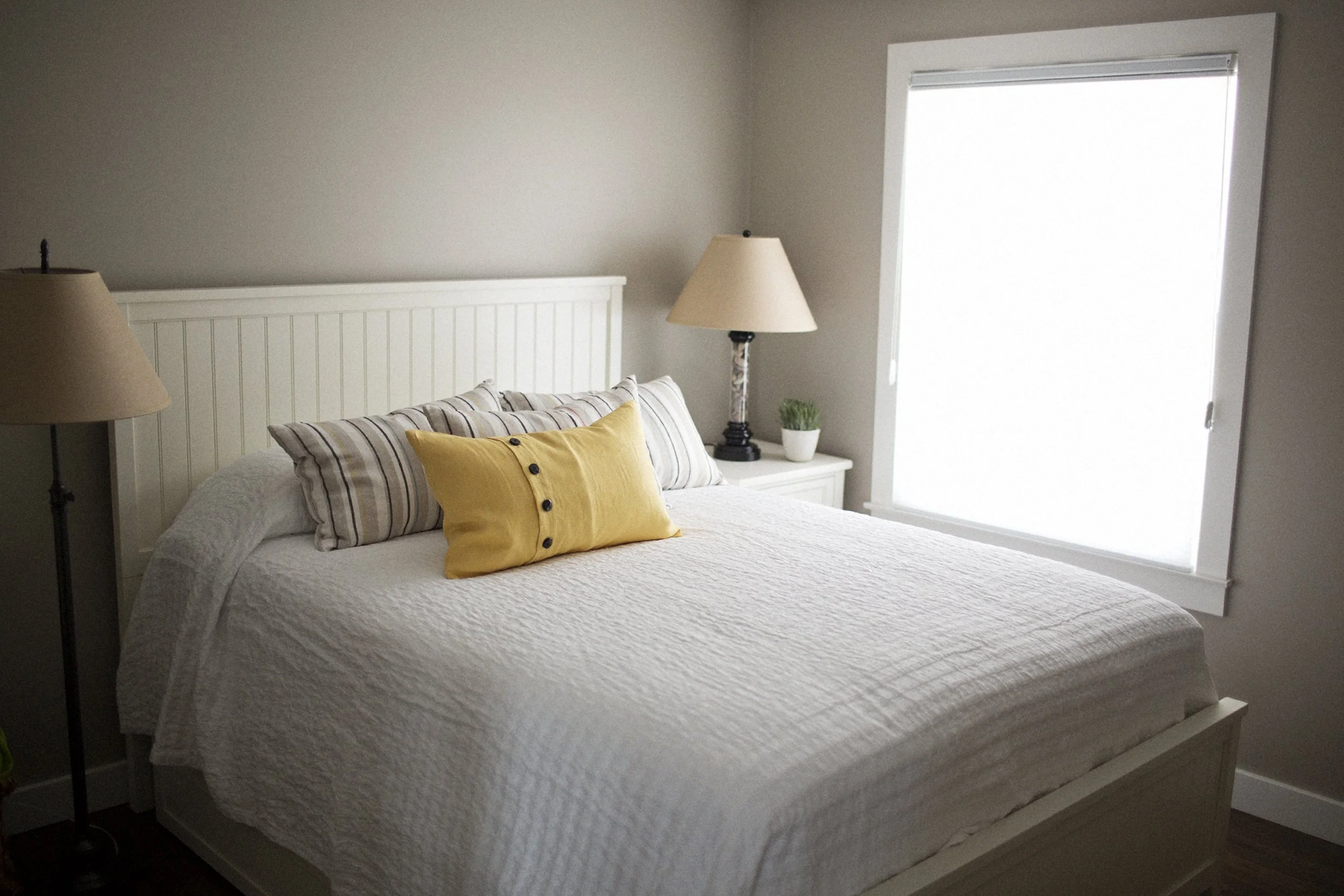 A neatly made bed with white bedding, striped and yellow pillows, in a bedroom with a window and two lamps on white nightstands.