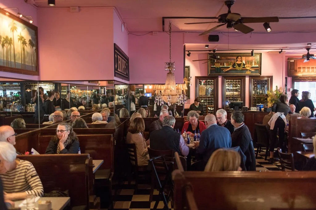 Indoor scene of a bustling restaurant or cafe with wooden booths, tables, and customers dining. The decor includes paintings, a chandelier, and a wine display. Waitstaff are serving and attending to patrons.