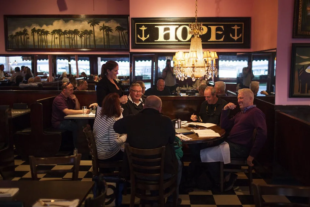 People dining in a restaurant with pink walls, a chandelier, and a sign that reads 'HOPE' with anchors on either side, large windows with checkered curtains, and paintings on the walls.