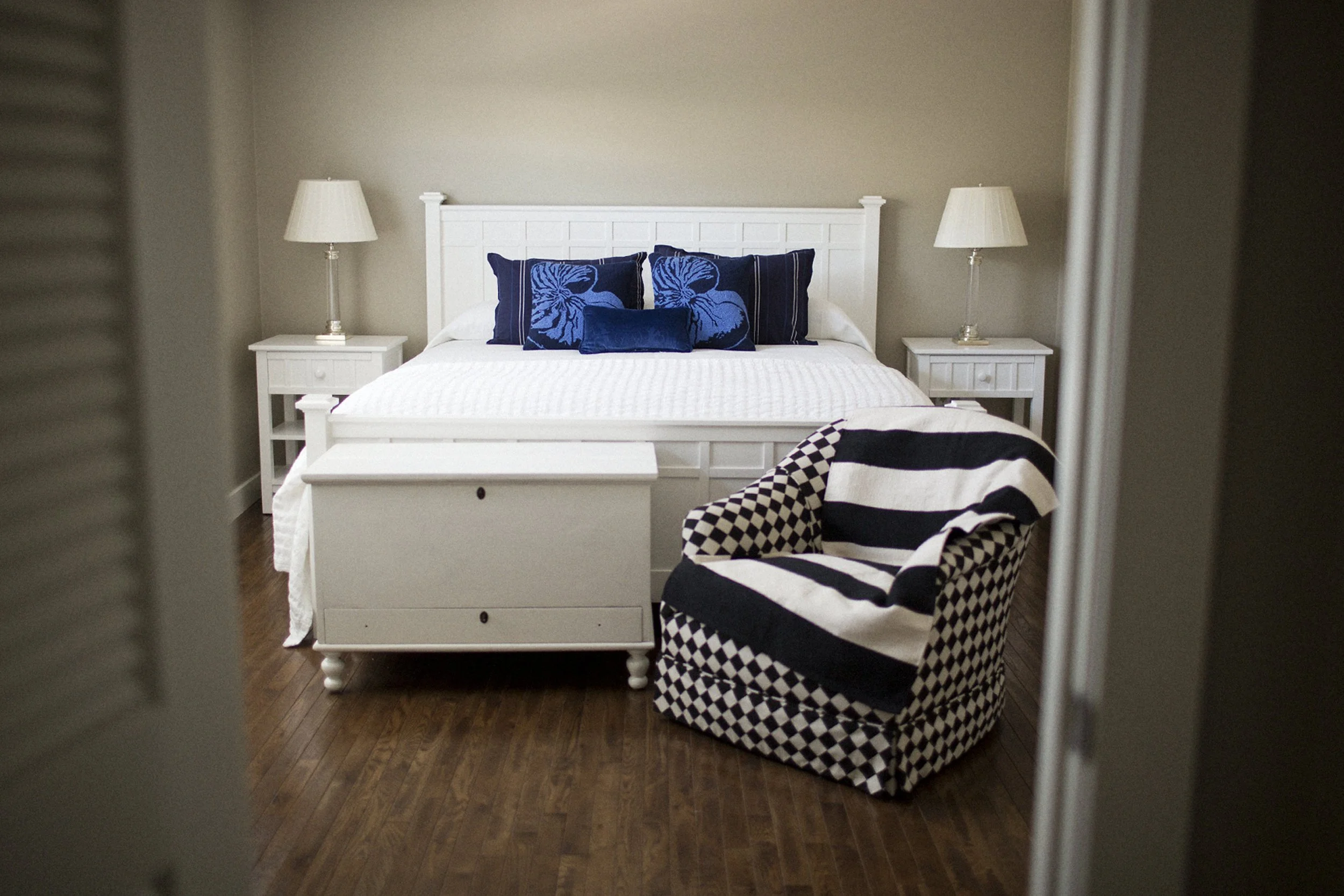 A bedroom with a white bed, dark blue pillows, white nightstands, two lamps, a black and white checkered armchair, and wooden flooring.