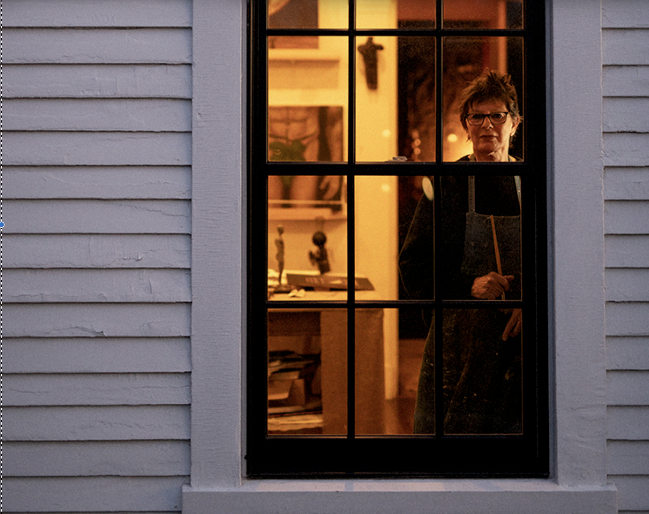 A woman standing inside a house, looking out the window. She is wearing glasses and a dark apron, holding a paintbrush. The interior is warmly lit, and there are shelves and decorative items visible in the background.