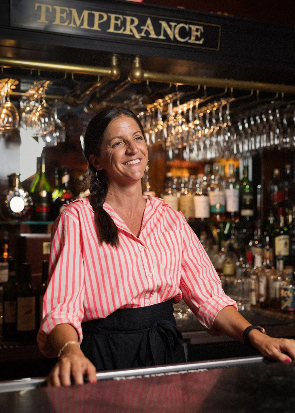 A smiling woman with dark hair in a braid, wearing a pink and white striped shirt, standing behind a bar with glasses hanging above her, bottles of liquor, and bar equipment in the background with a "Temperance" sign overhead.