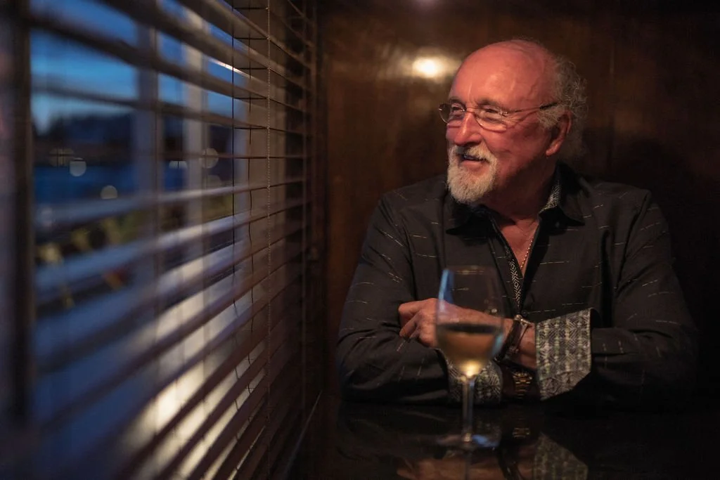 An elderly man with glasses and a beard, dressed in a dark shirt, sitting at a table with a glass of white wine, smiling and looking outside through window blinds in a dimly lit setting.