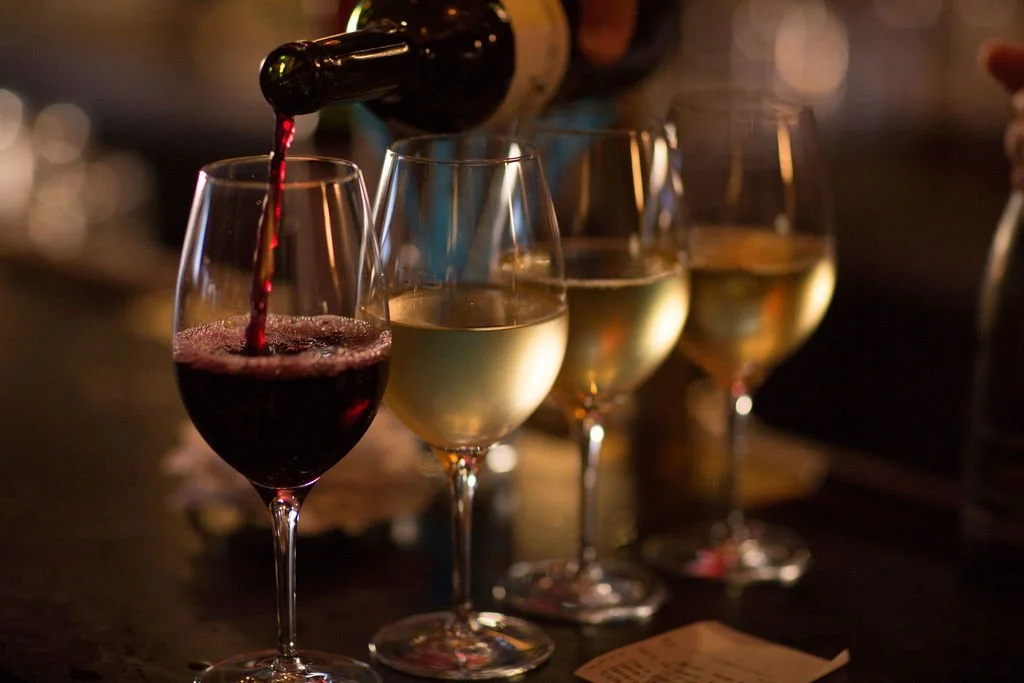 A hand pouring red wine into a glass, with three other glasses of white wine in the background on a dark bar counter.