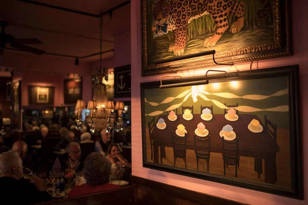 Dimly lit restaurant with people dining, featuring paintings of cows and cupcakes on the wall.