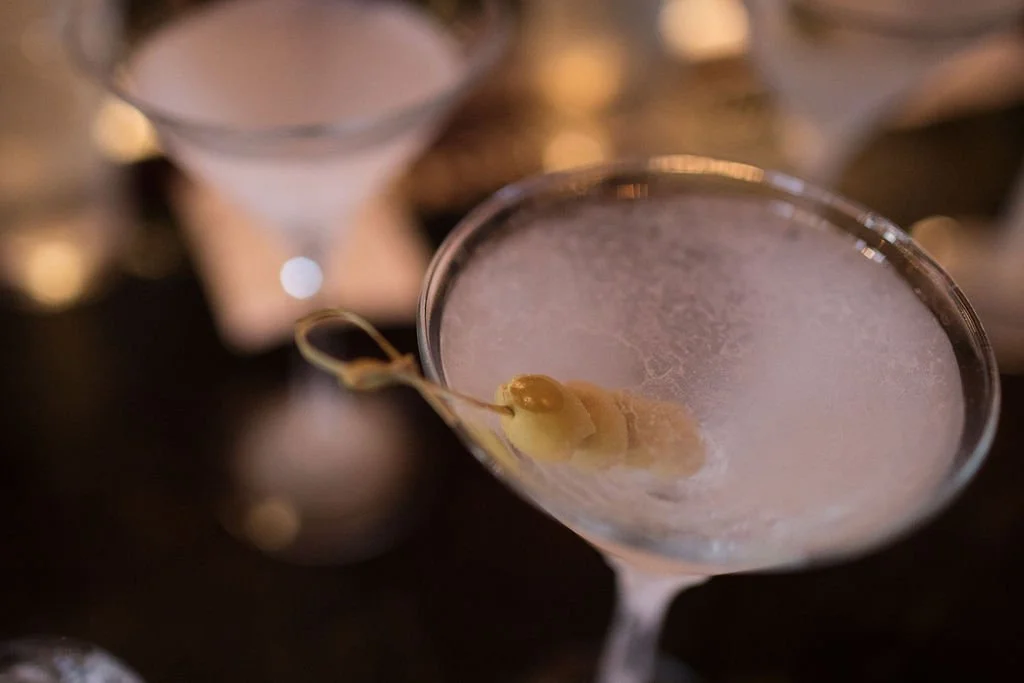 Close-up of a cocktail glass with a frosted surface, garnished with an olive on a skewer, on a dark bar counter.