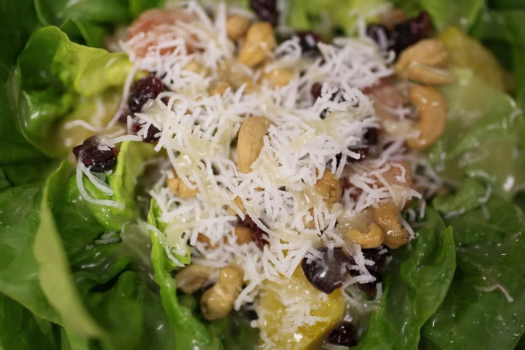 Close-up of a salad with green lettuce, shredded cheese, dried cranberries, walnuts, and chopped pickles.