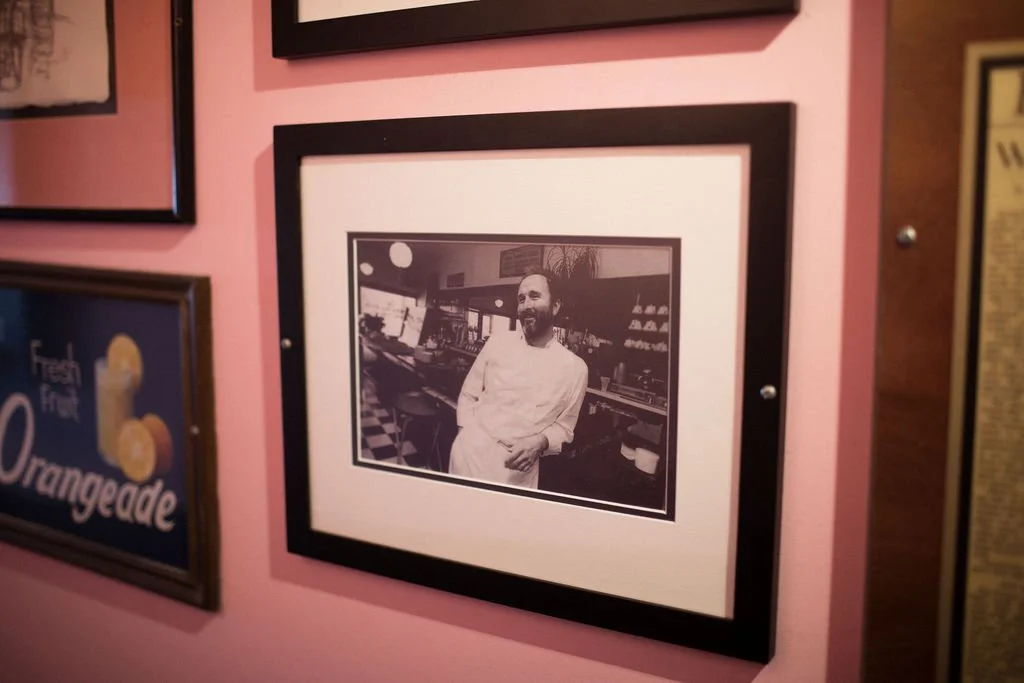 Black and white photograph of a man with a beard wearing a white chef's coat, standing in a restaurant or bar, with background decor including shelves and windows, framed on a pink wall.