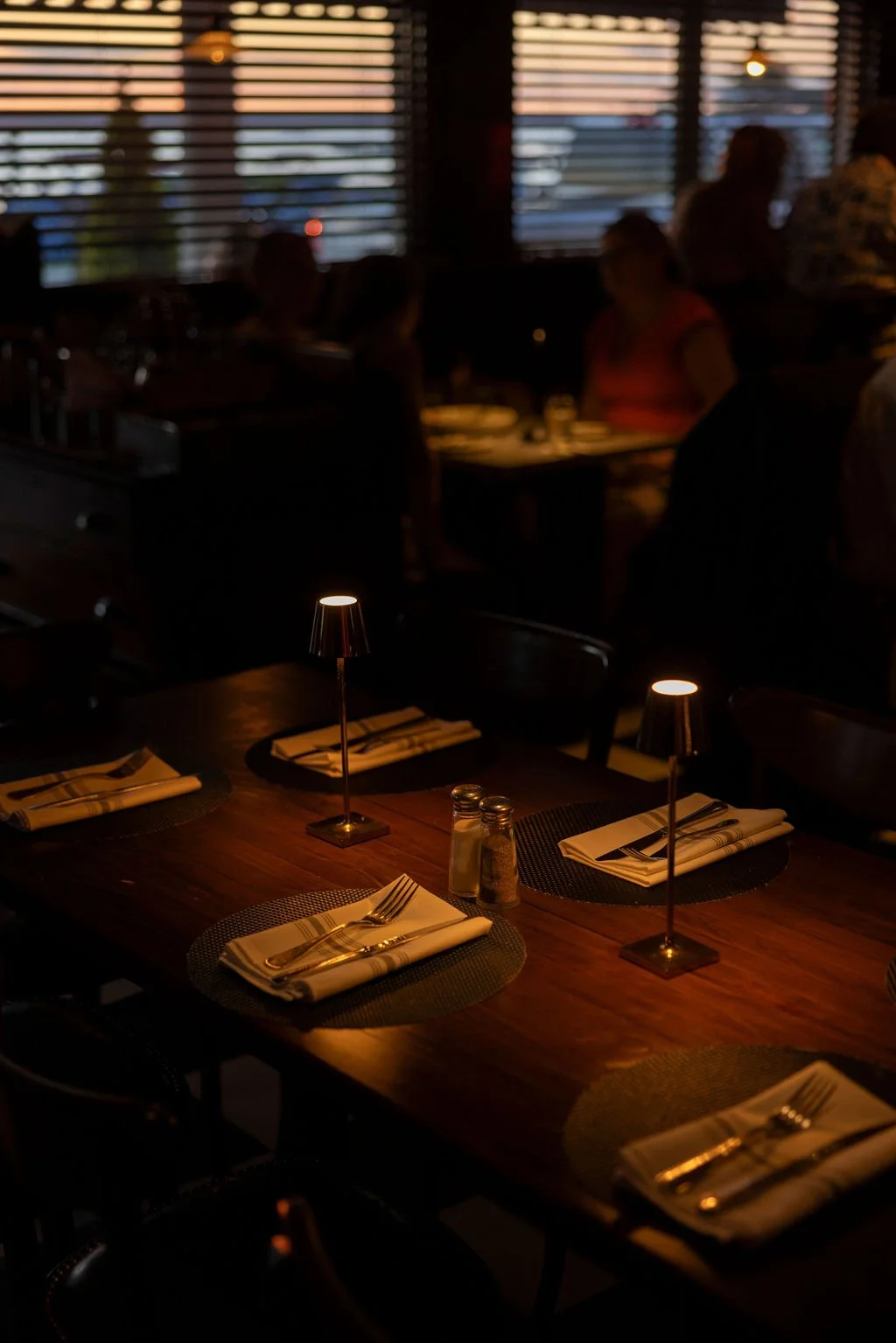 Empty dining table set with napkins, forks, knives, salt and pepper shakers, small lamps, in a dimly lit restaurant, with blurred diners and window blinds in the background.
