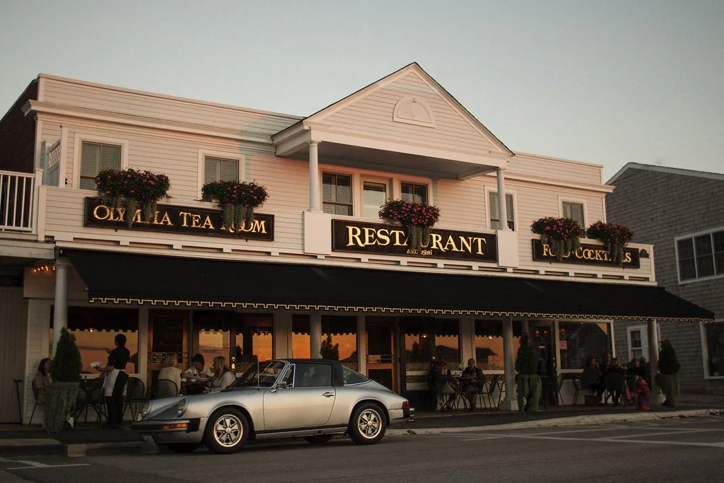 White multi-story building housing a restaurant, tea museum, and food court, with outdoor seating and people dining, a vintage silver car parked in front, and decorative flower boxes on the upper balcony.